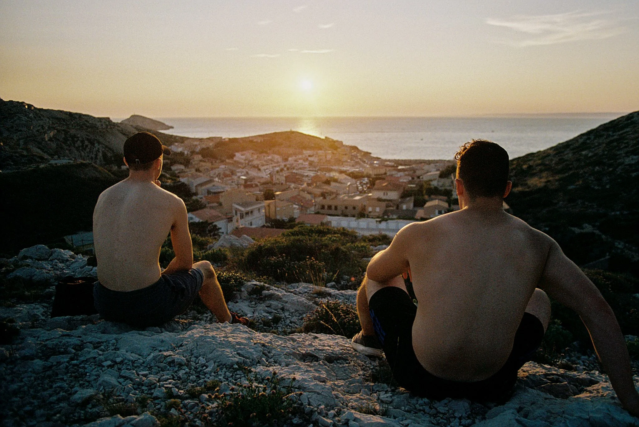 Two shirtless young men sitting on rocks overlooking a coastal town at sunset with the ocean and sky in the background.