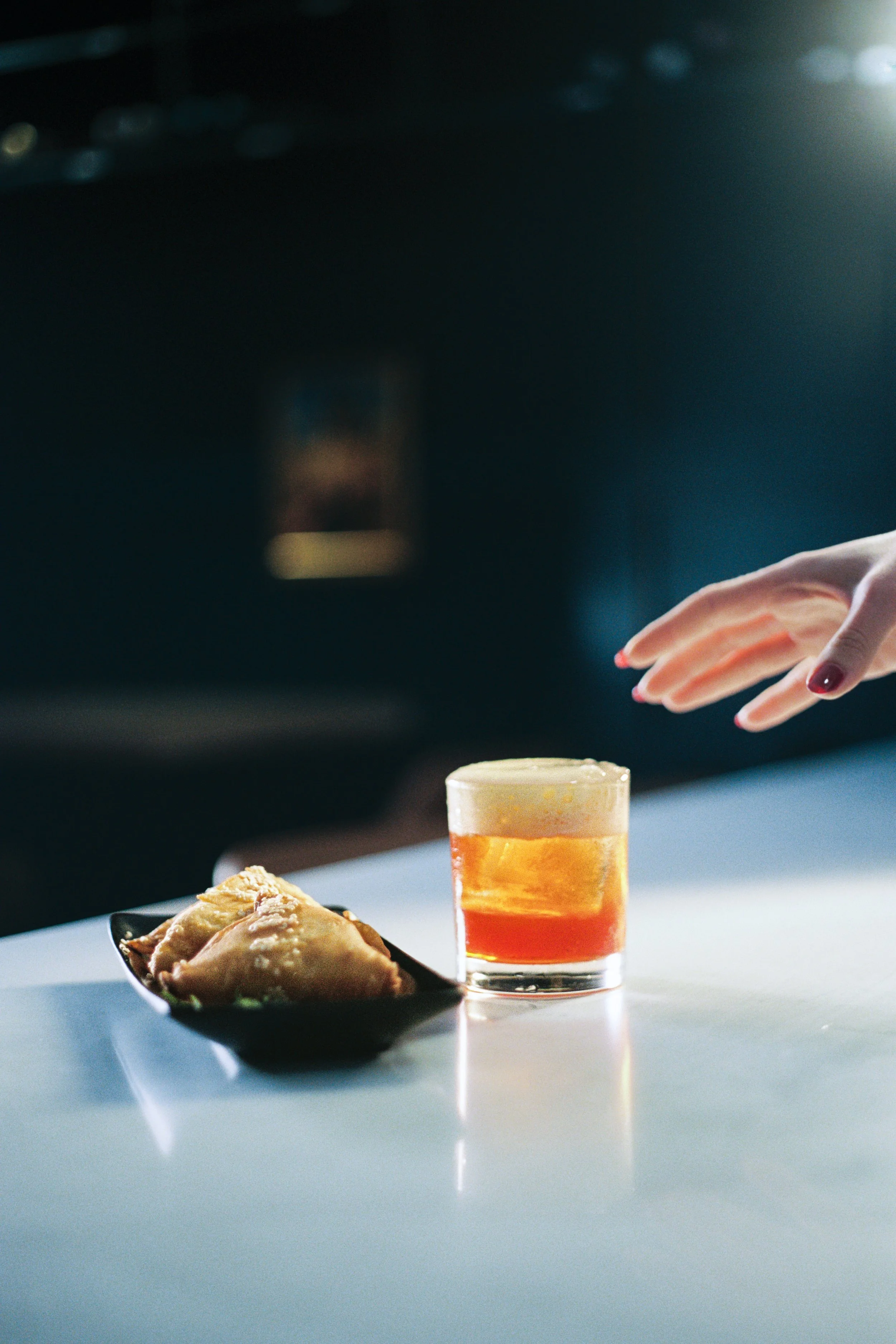 Close-up of a partially eaten slice of pizza on a black plate, a glass of layered orange and yellow beverage, and a hand with manicured nails reaching towards the food on a white table, with a dark background.