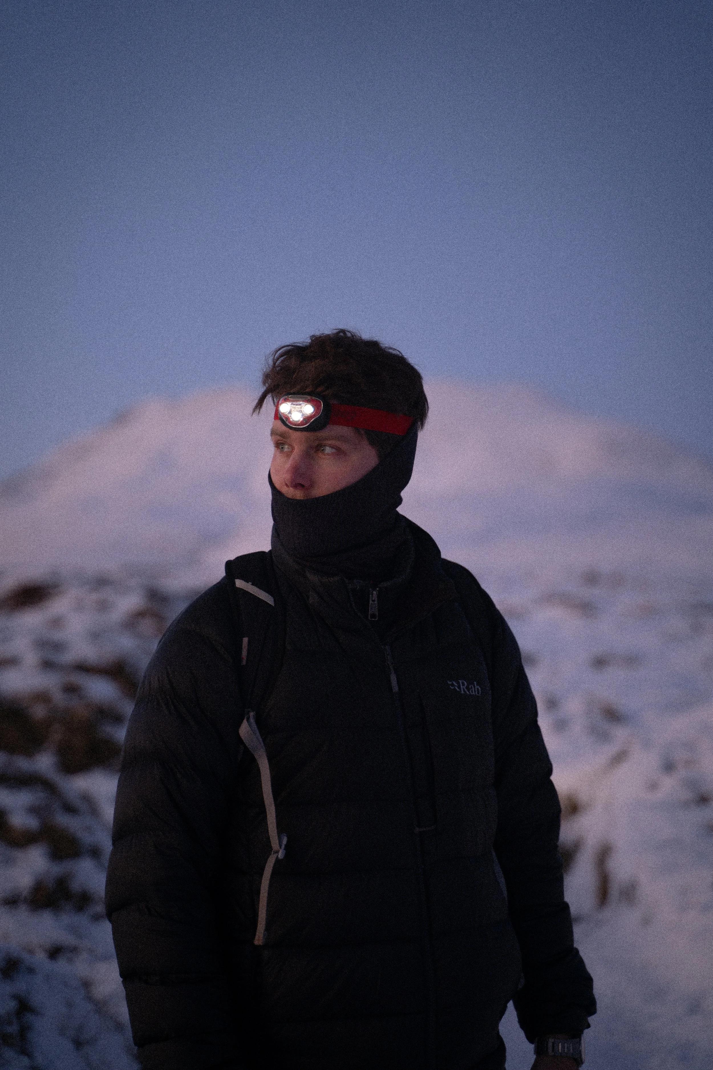 A man outdoors in a snowy landscape during twilight or early evening, wearing a black jacket, a headlamp, and a neck gaiter.