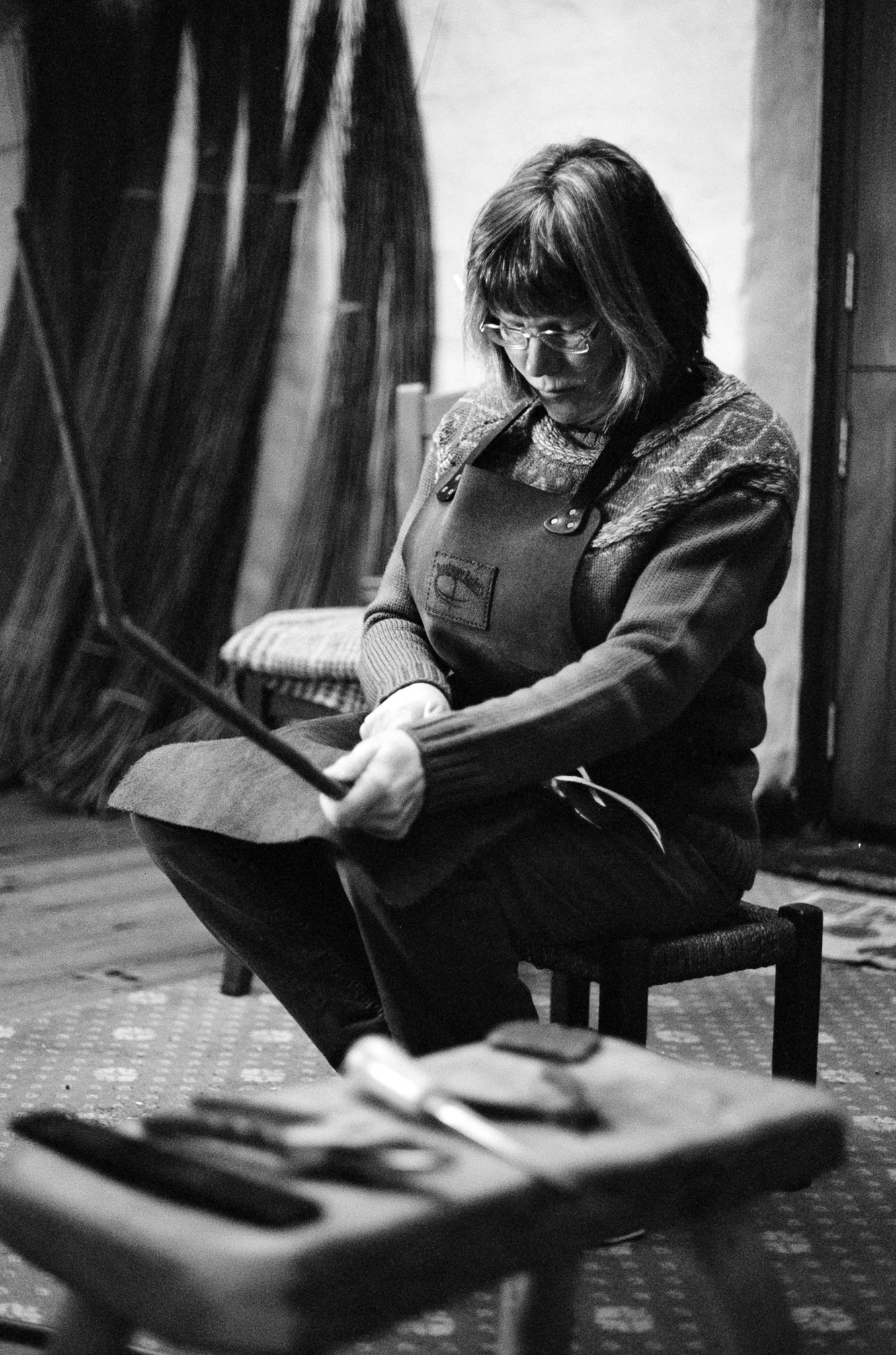A woman with glasses and shoulder-length hair wearing an apron over a patterned sweater, sitting on a chair with her head bowed and eyes closed, in an indoor setting with wooden floors and a textured wall.