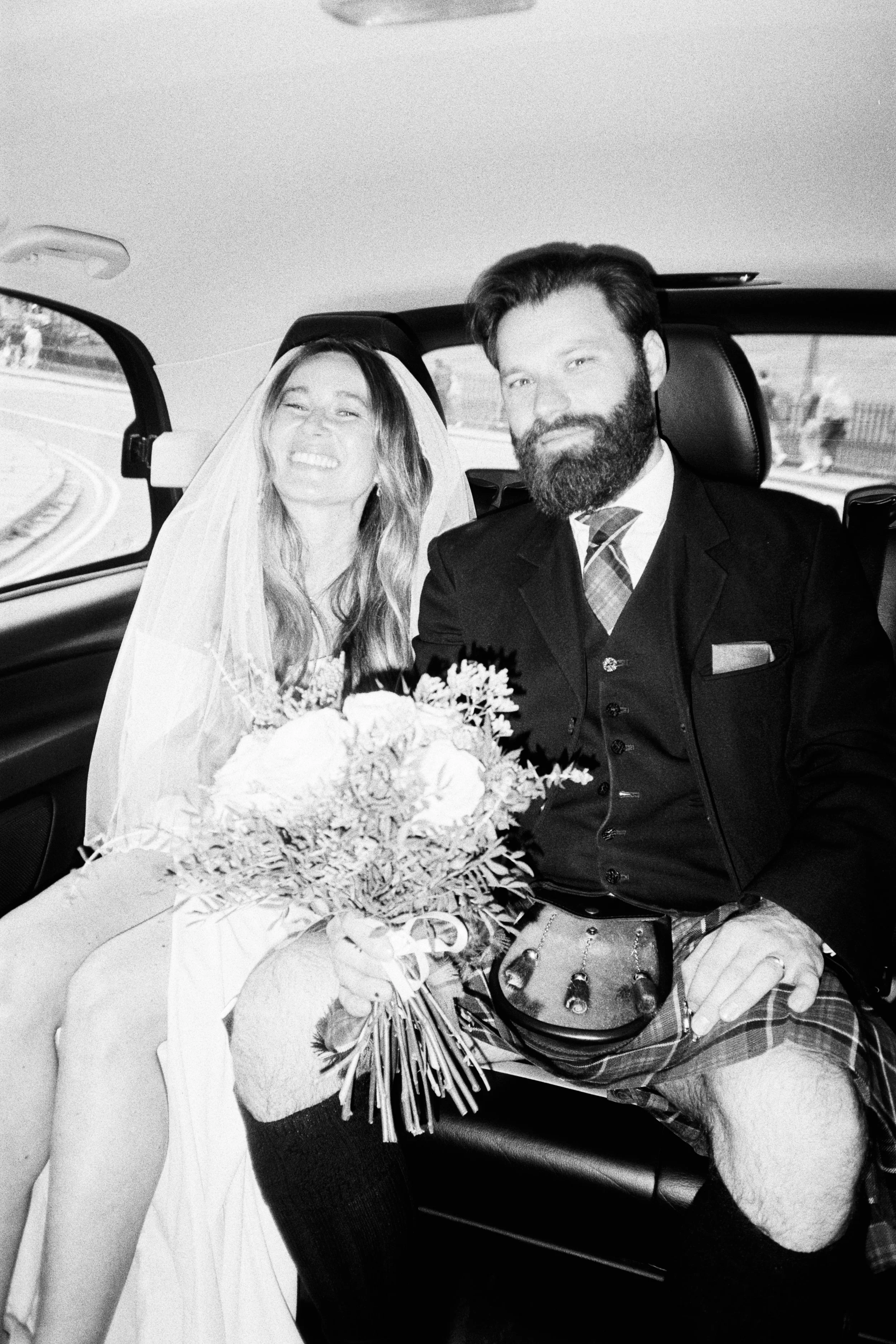 Black and white photo of a bride and groom sitting inside a vehicle. The bride is smiling, wearing a wedding veil, and holding a bouquet. The groom has a beard, is dressed in a suit with a patterned tie, and is seated beside her.