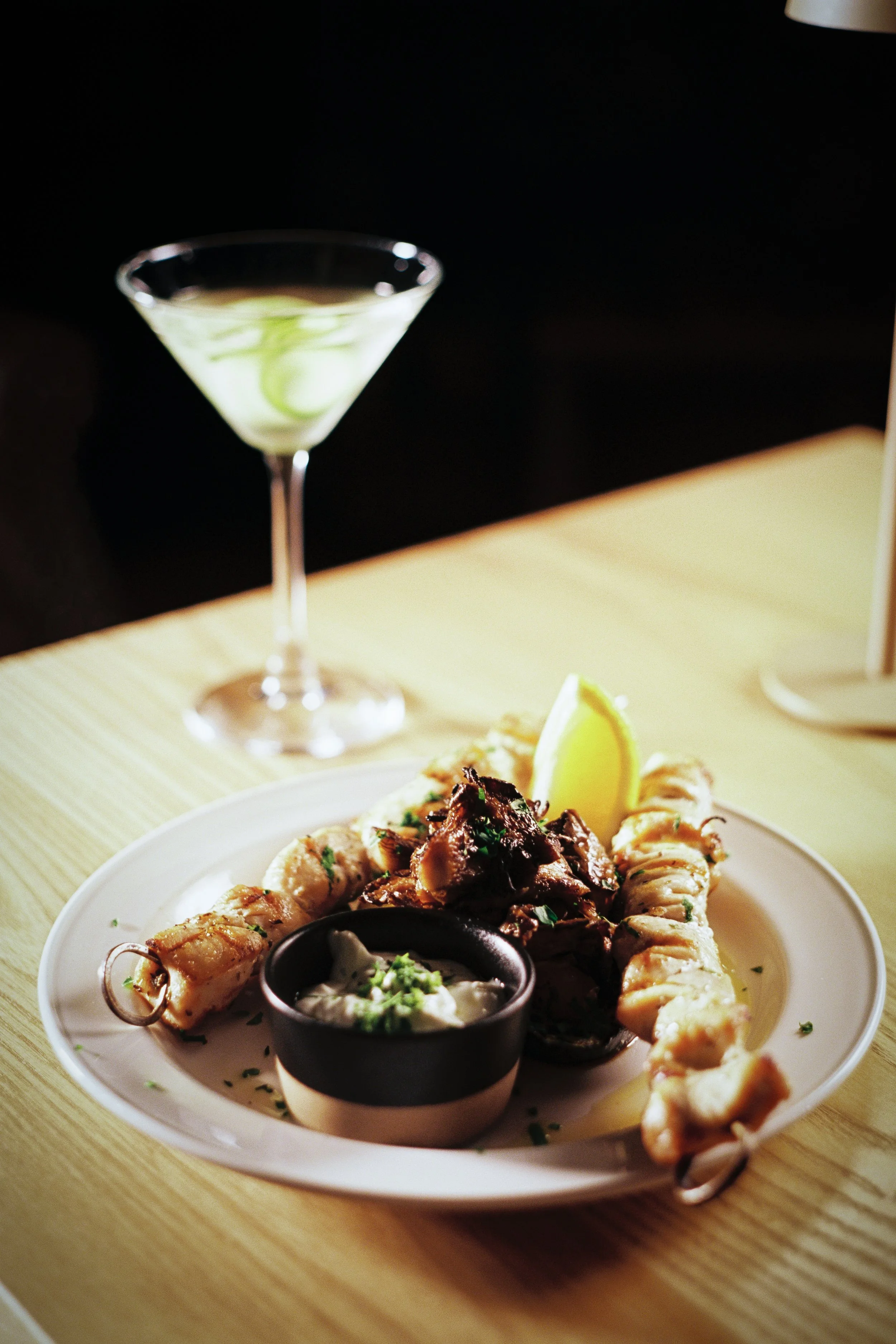 A plate of assorted seafood, including shrimp, topped with a lemon wedge and served with garnishes, with a martini glass containing a lime twist in the background.