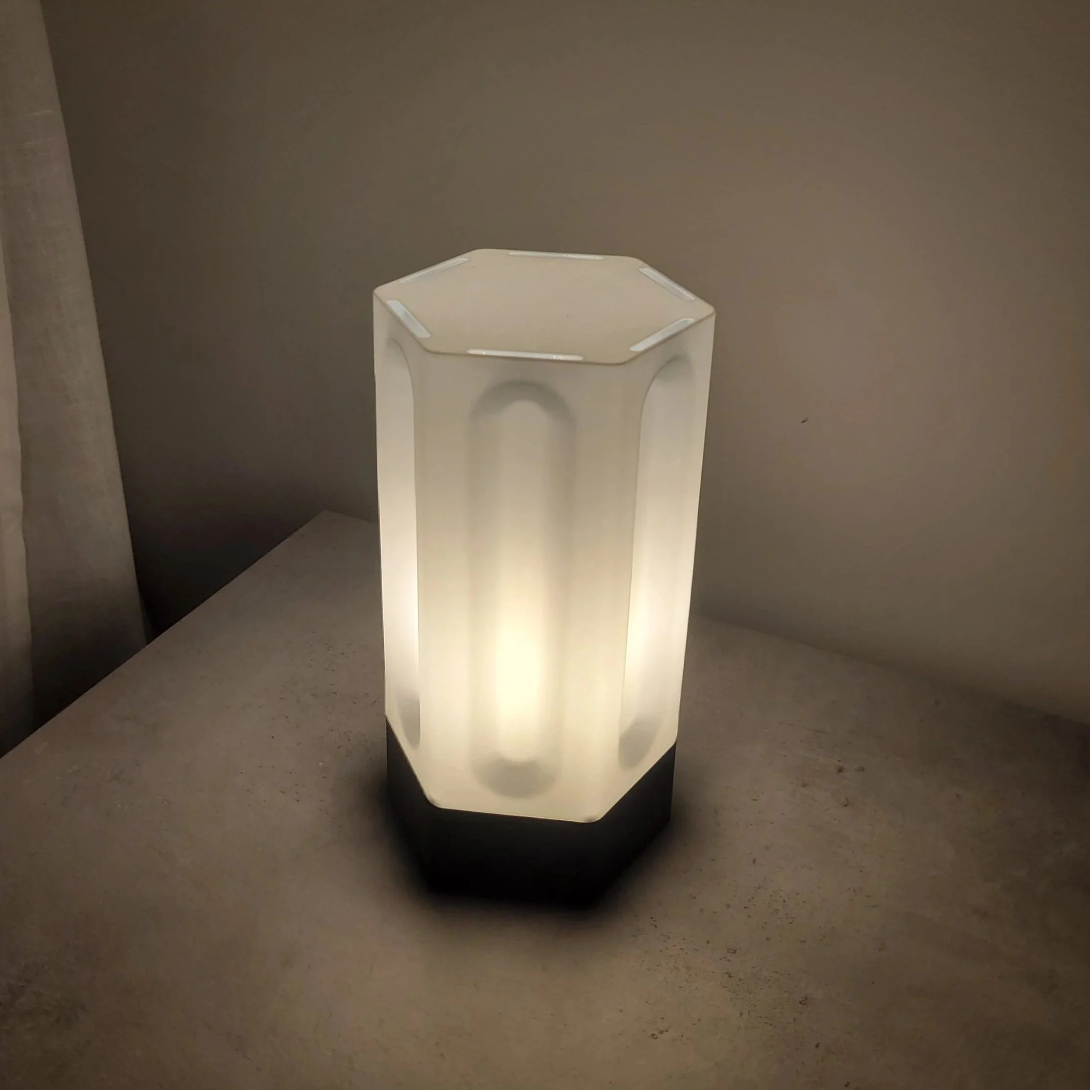 A white pedestal with a hexagonal shape and a textured surface, placed on a beige floor against a plain beige wall.