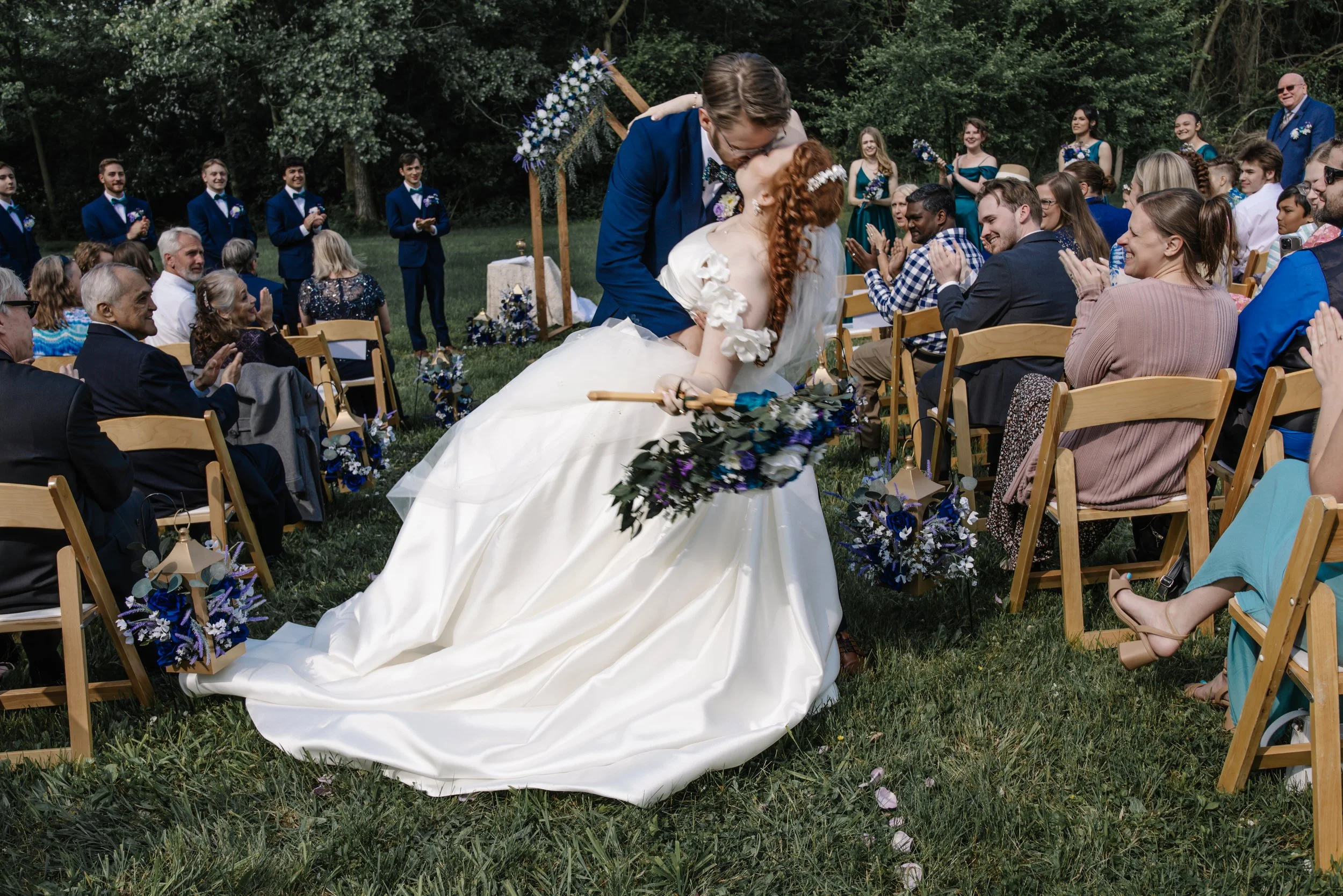 Couple kissing at an outdoor wedding ceremony, with guests seated on wooden chairs and an arch decorated with flowers in the background.