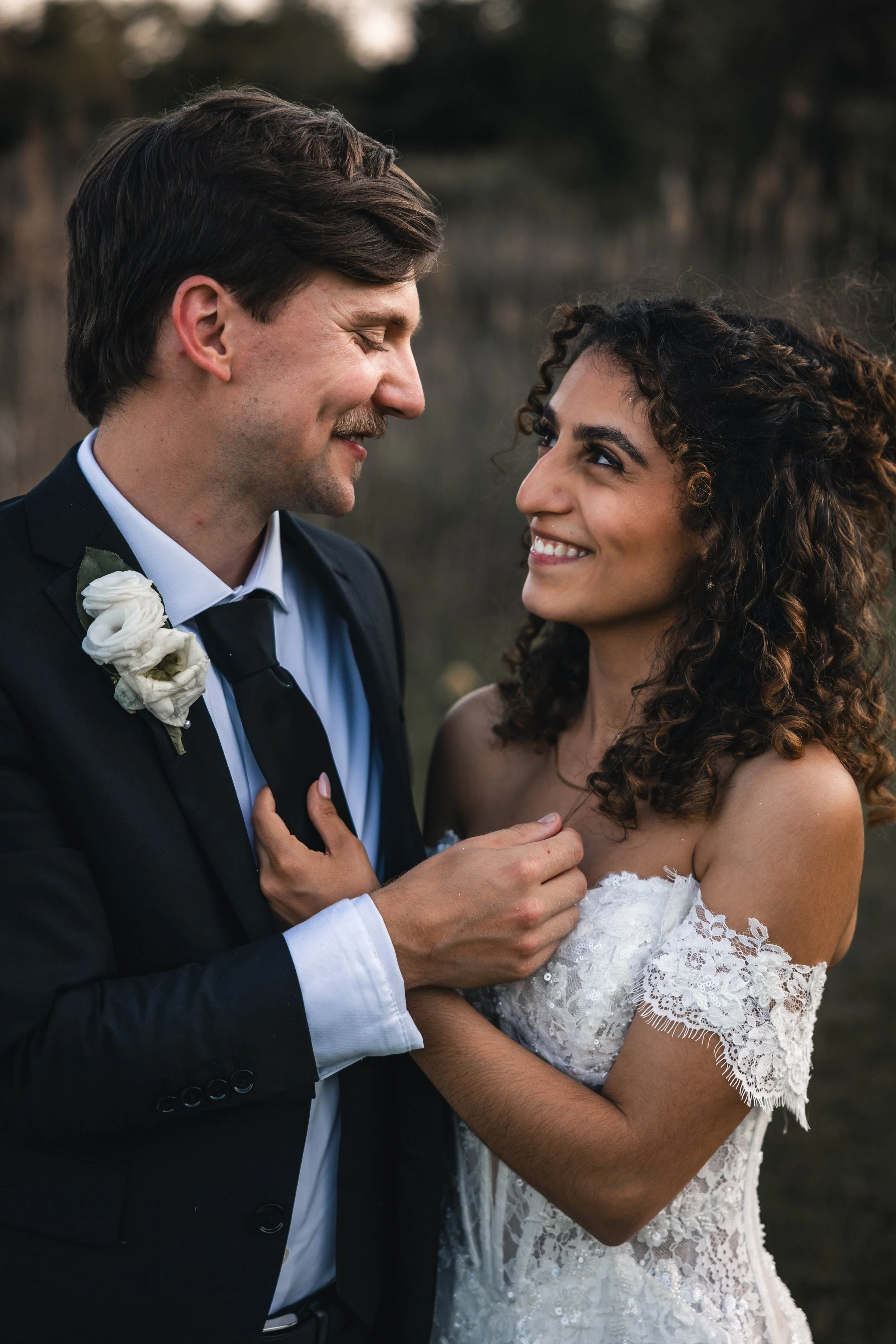 A man in a black suit with a white shirt and boutonniere, and a woman in a white lace wedding dress, sharing a tender moment outdoors with bodies close and smiling.