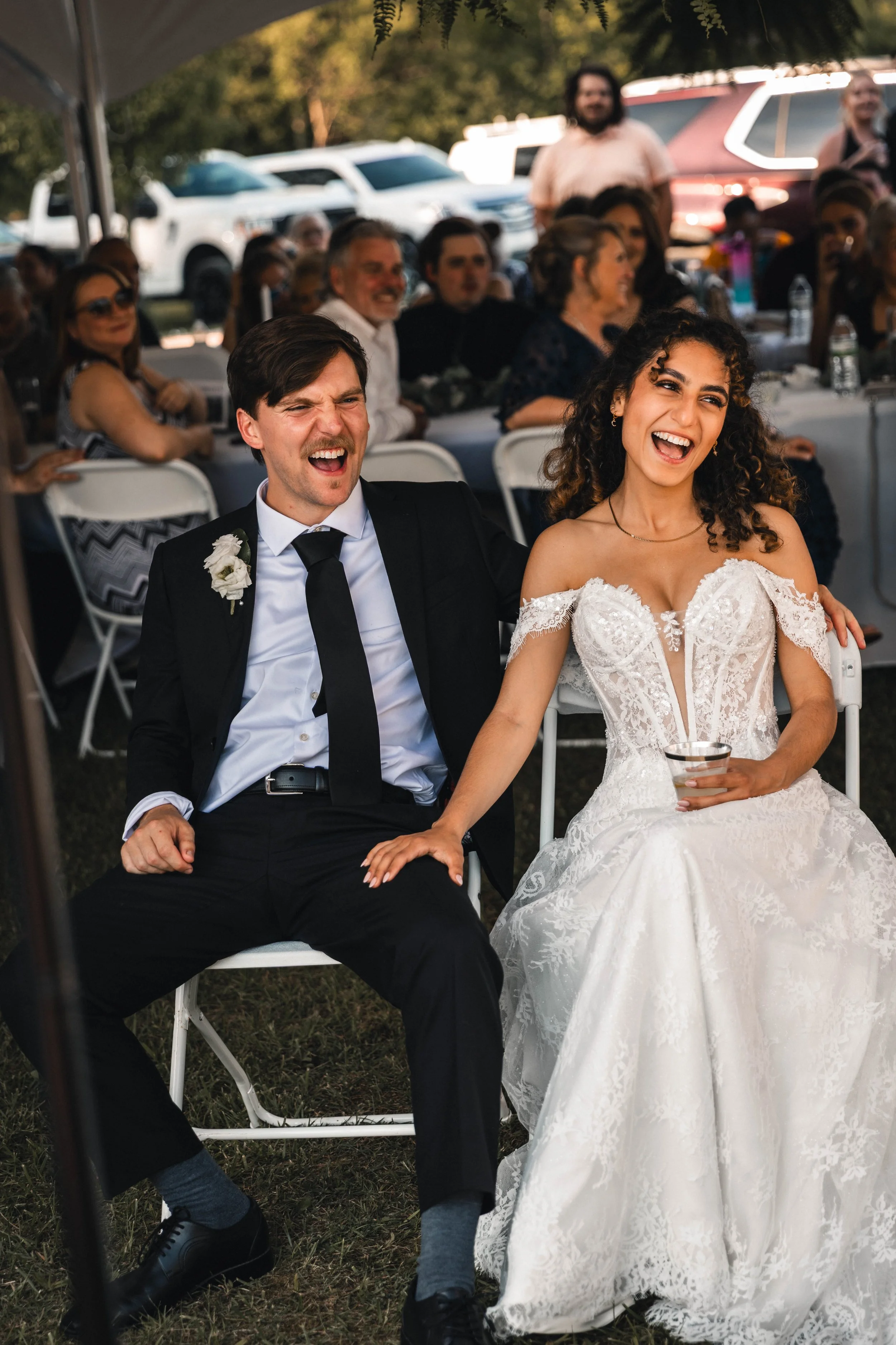 A joyful bride and groom sitting together at their wedding reception outdoors, with wedding guests seated around them under a tent.