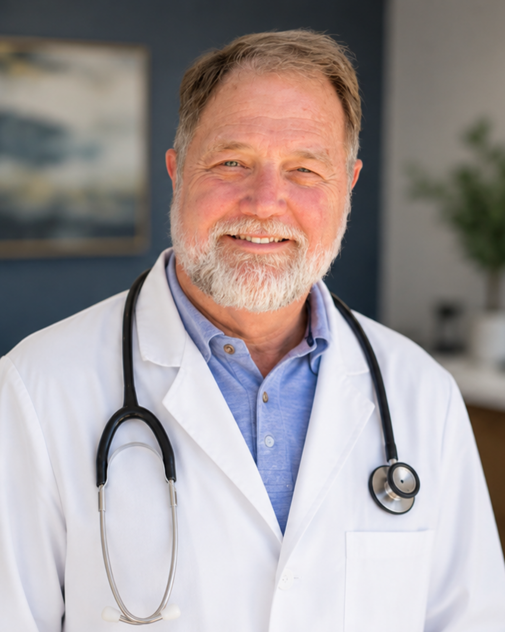 A senior male doctor with a beard and mustache, wearing a white lab coat and a light blue shirt, with a stethoscope around his neck, smiling.