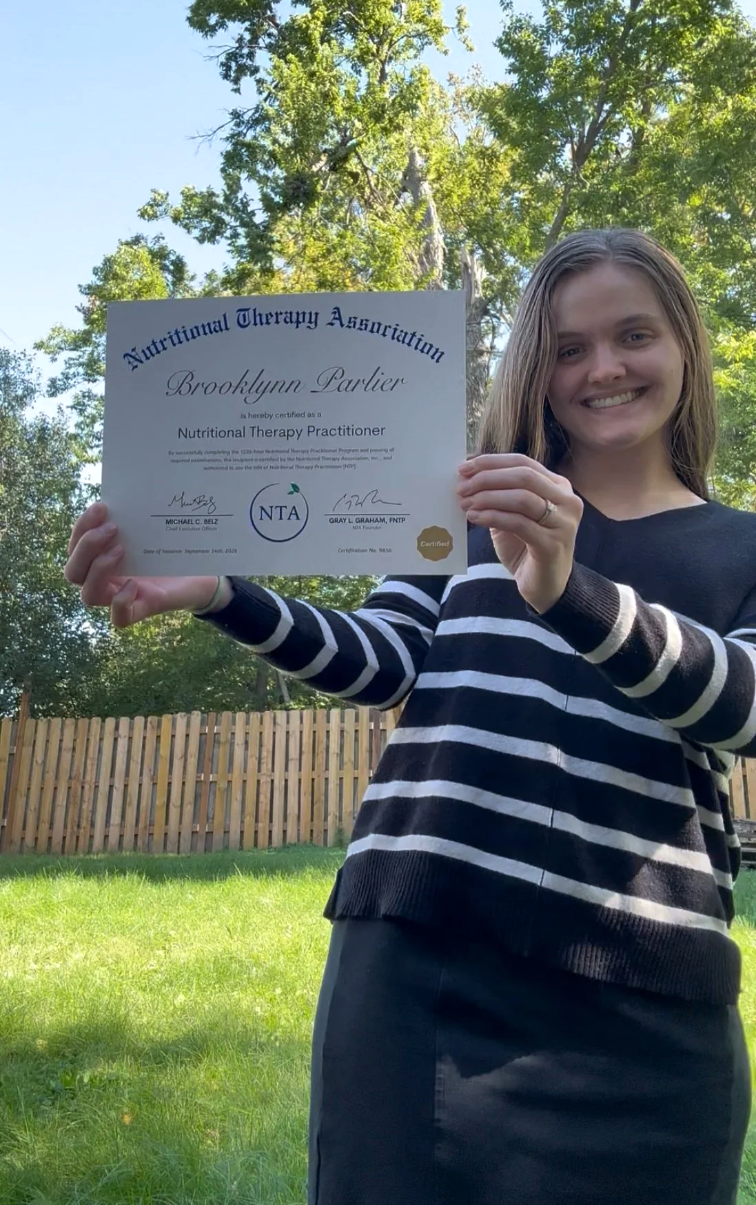 A woman smiling outdoors holding a certificate for Nutritional Therapy Practitioner from Nutritional Therapy Association.