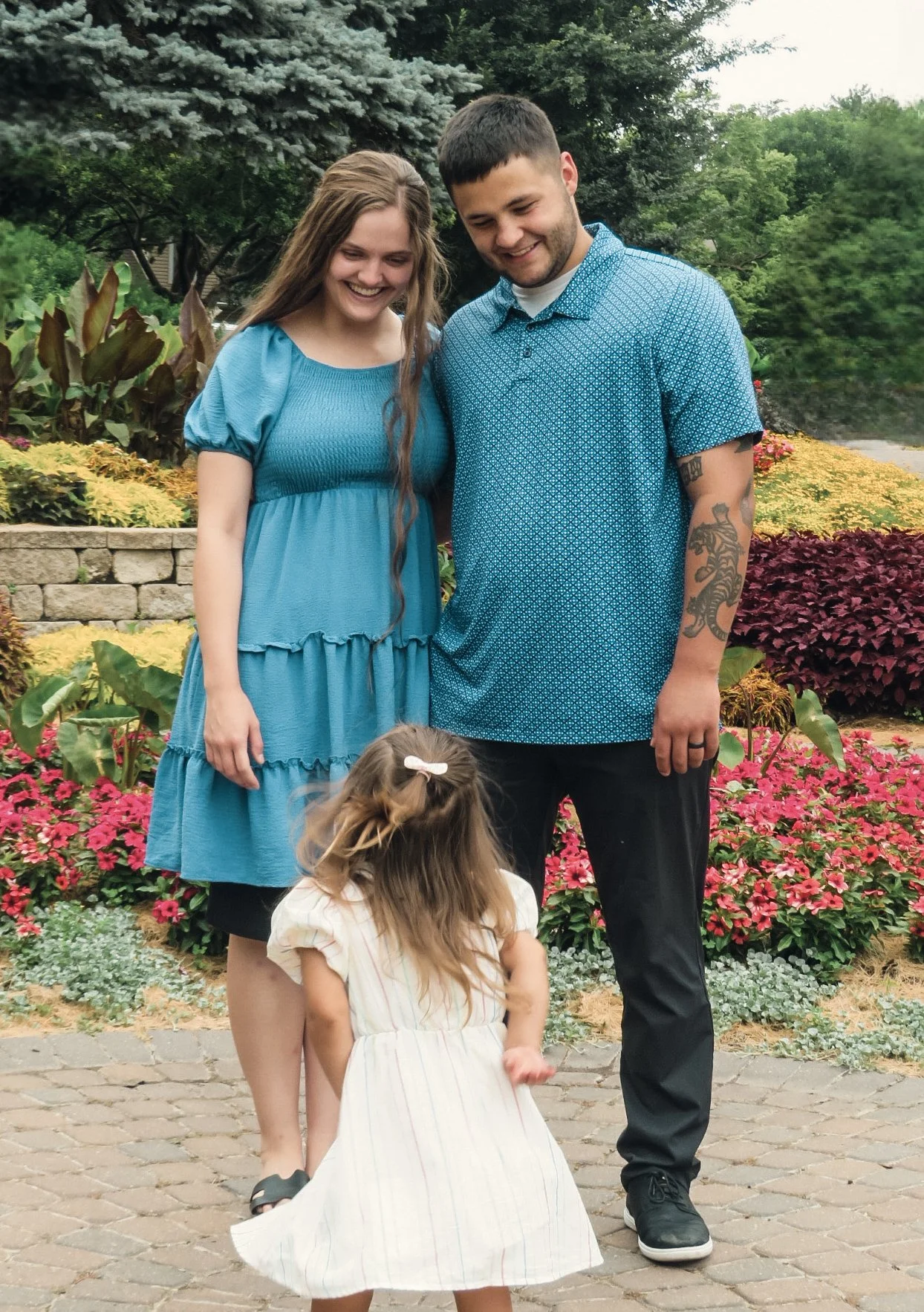 A family of four, including a woman in a blue dress, a man in a blue patterned shirt with tattoos, and two young girls, one with a bow in her hair, standing outdoors on a brick pathway in front of a colorful flower garden with trees.
