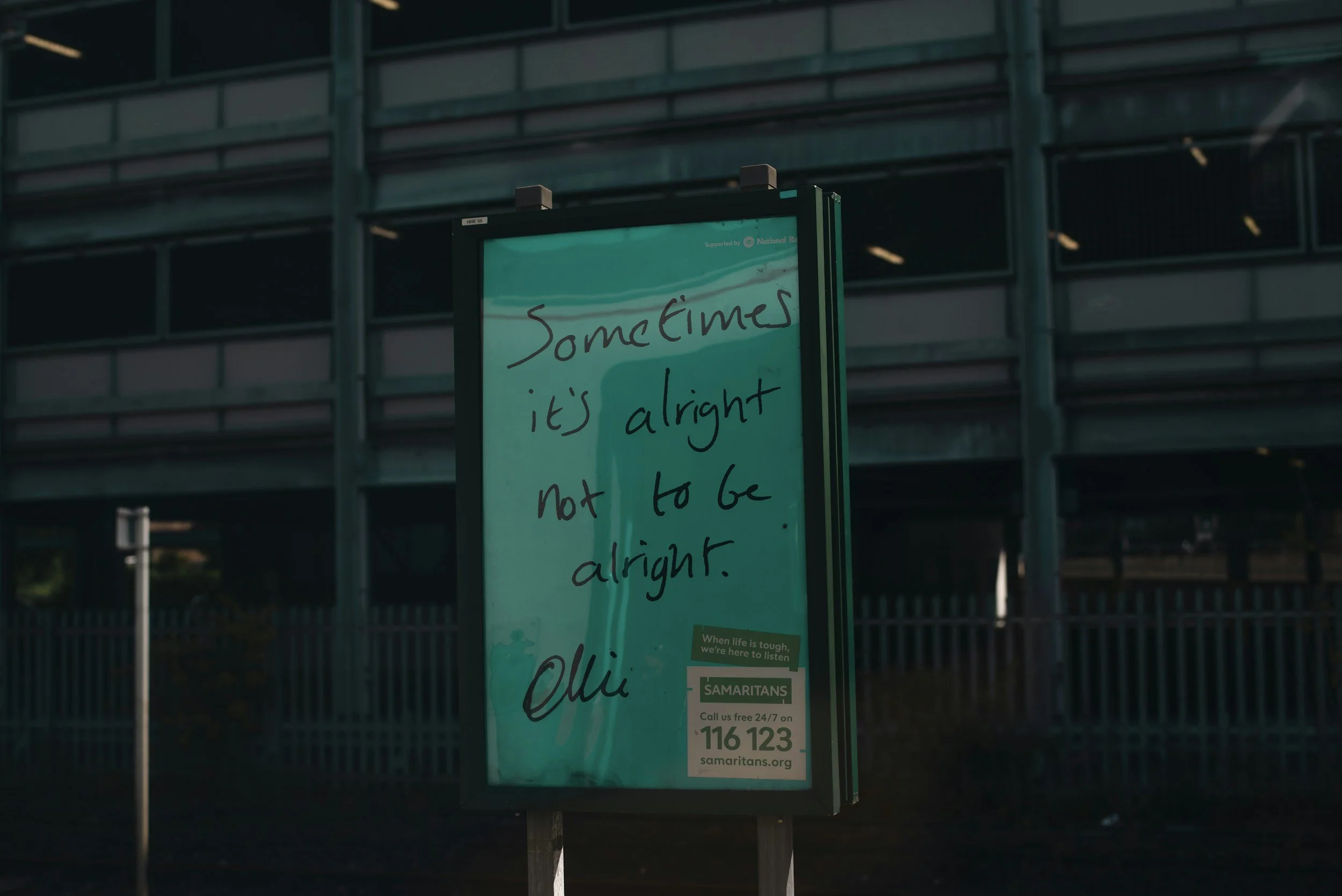 A digital informational sign with handwritten text that reads 'Sometimes it's alright not to be alright,' positioned outdoors near a modern building with green-tinted windows.