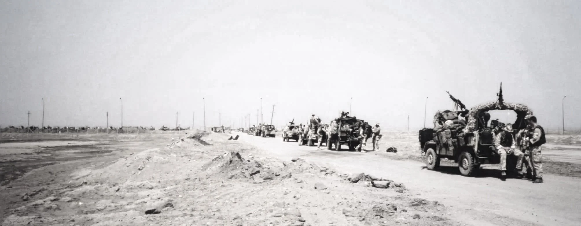 Black-and-white photo of soldiers and military vehicles traveling on a dirt road in a desert landscape with sparse power lines in the distance.