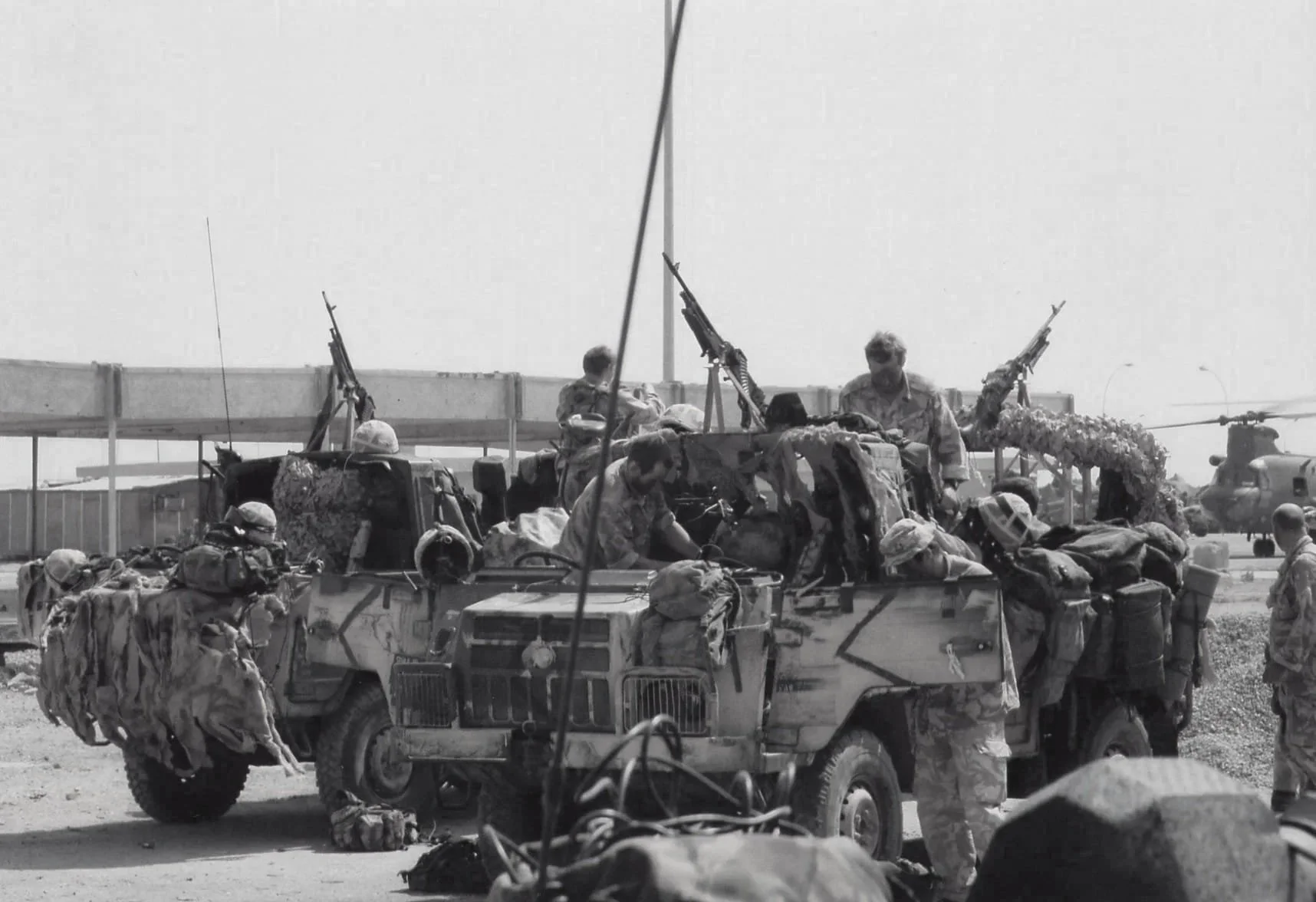Black and white photo of military soldiers with a mounted military vehicle, soldiers in uniform unloading supplies, and helicopters in the background.
