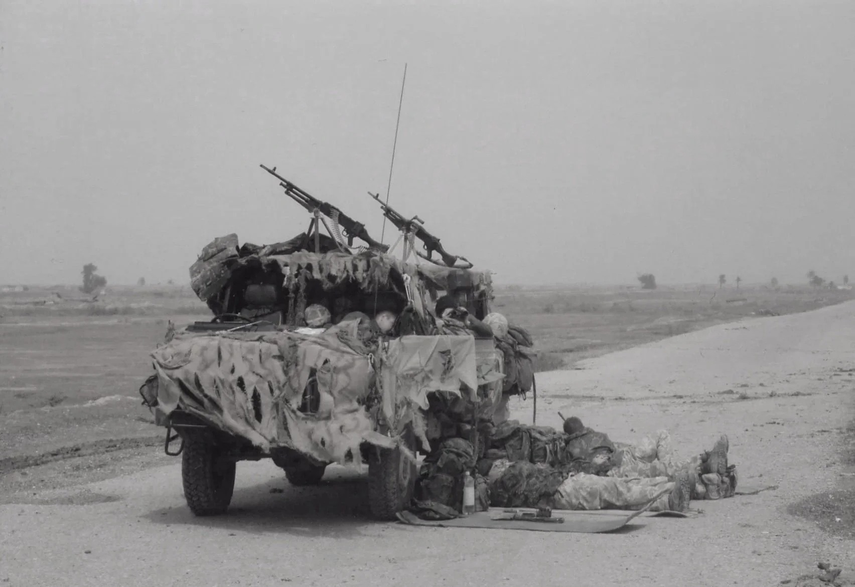 Soldiers resting beside a military vehicle with two mounted machine guns in a desert landscape.