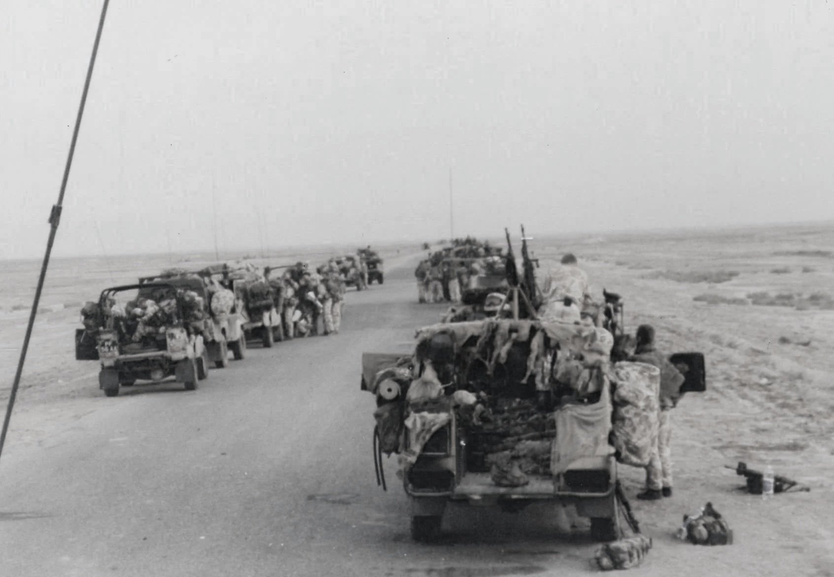 Black and white photo of soldiers walking or standing near military vehicles on a flat, open desert landscape with a clear horizon and no visible buildings or trees.