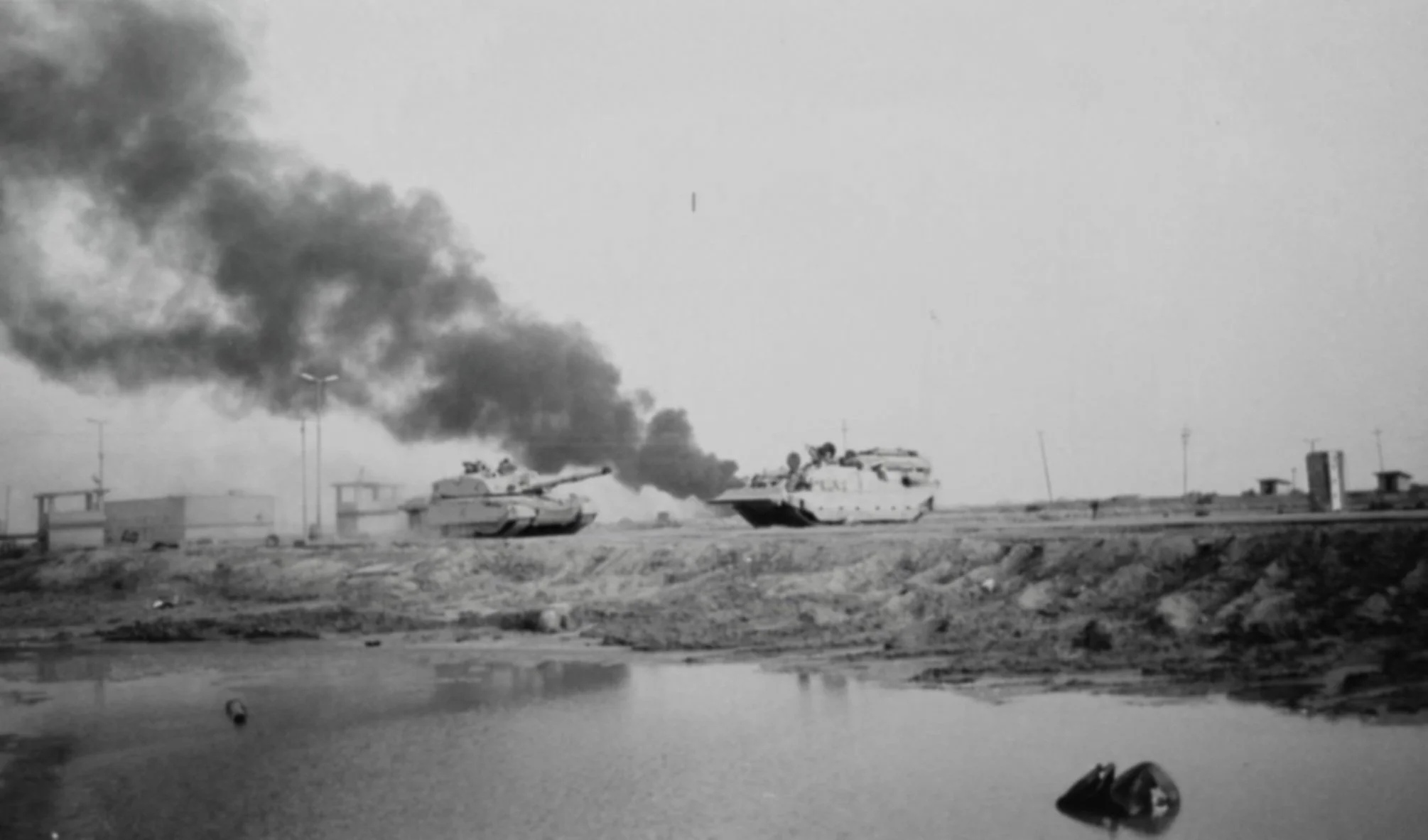 Black and white photo of two tanks on a flat open terrain with smoke rising from one of them, a body of water in the foreground, and a few small buildings or structures in the background.