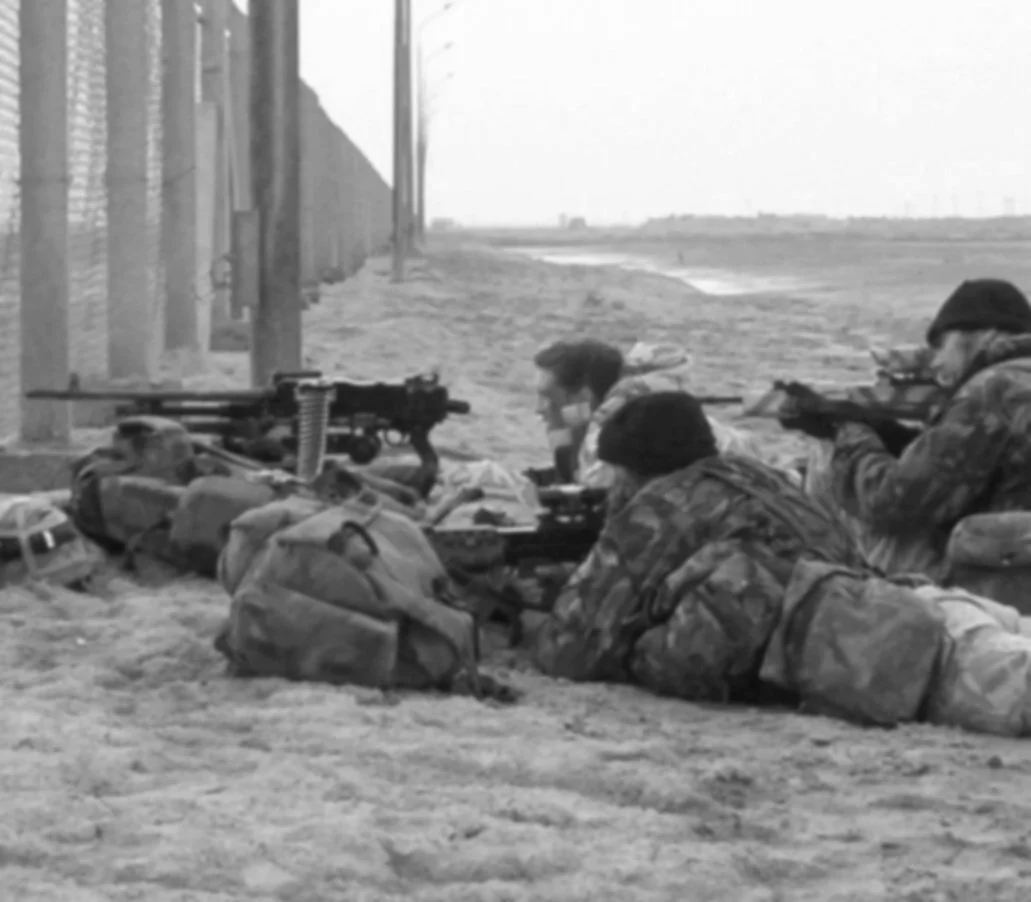 Three soldiers in camouflage uniforms lying on the ground aiming rifles through a barricade at a pay station on a beach.