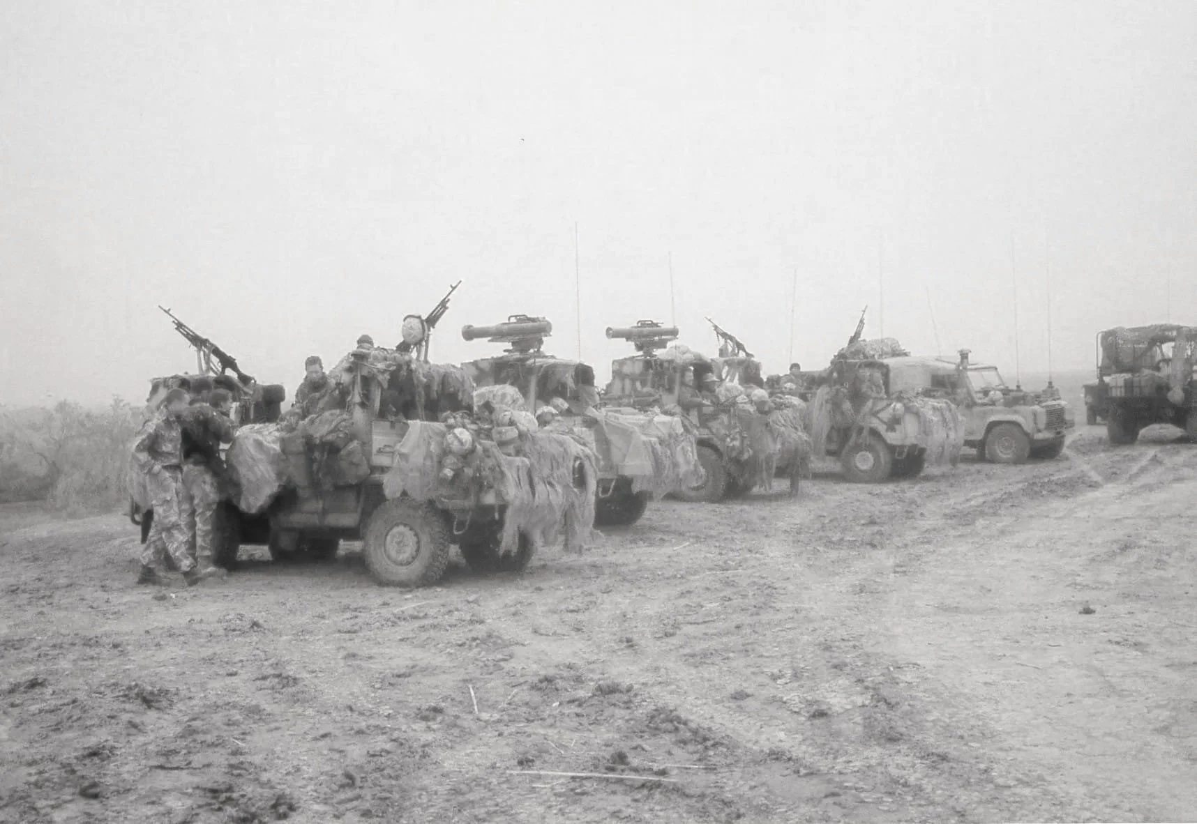 Black and white photo of military troops around several armored vehicles with mounted guns, on a dirt road in a rural area.