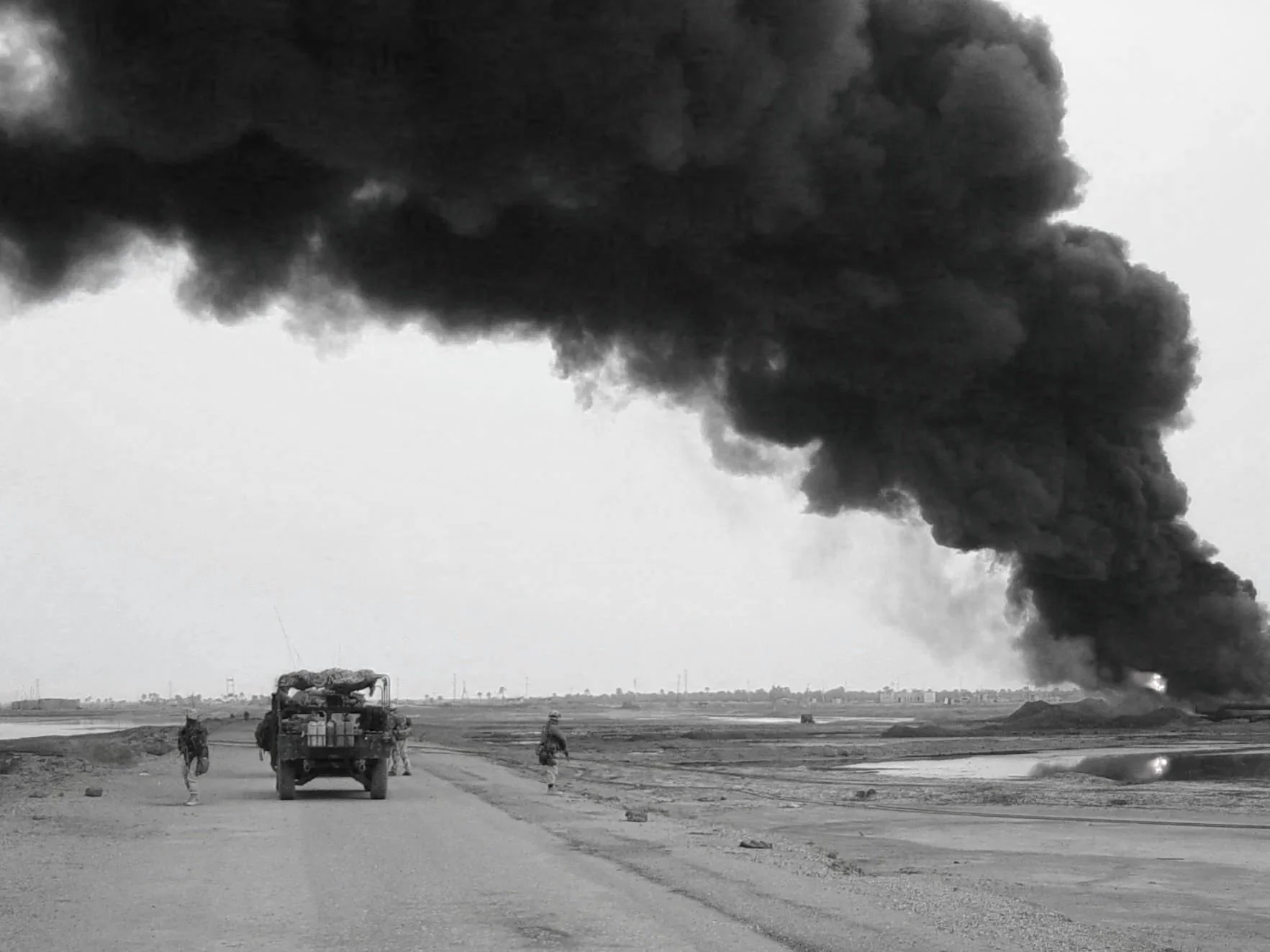A landscape with a large plume of black smoke rising from the ground, possibly from a fire or explosion. Several people and vehicles are visible on the dirt road in the foreground, with the scene appearing to be in an open, possibly industrial or con