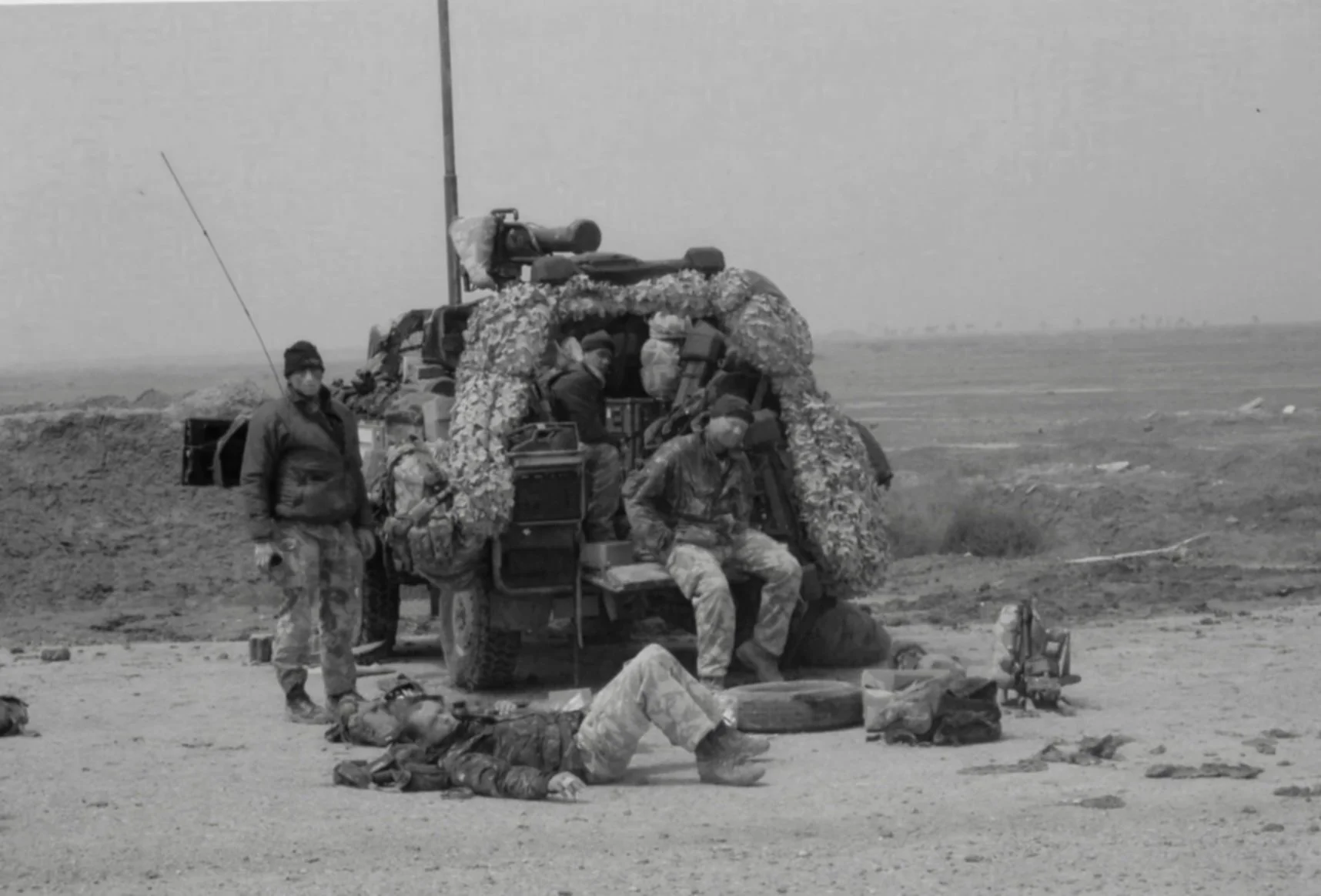 Black and white photograph of soldiers resting in a barren field with a military vehicle behind them; some soldiers are sitting, others standing or lying on the ground, surrounded by equipment and supplies.