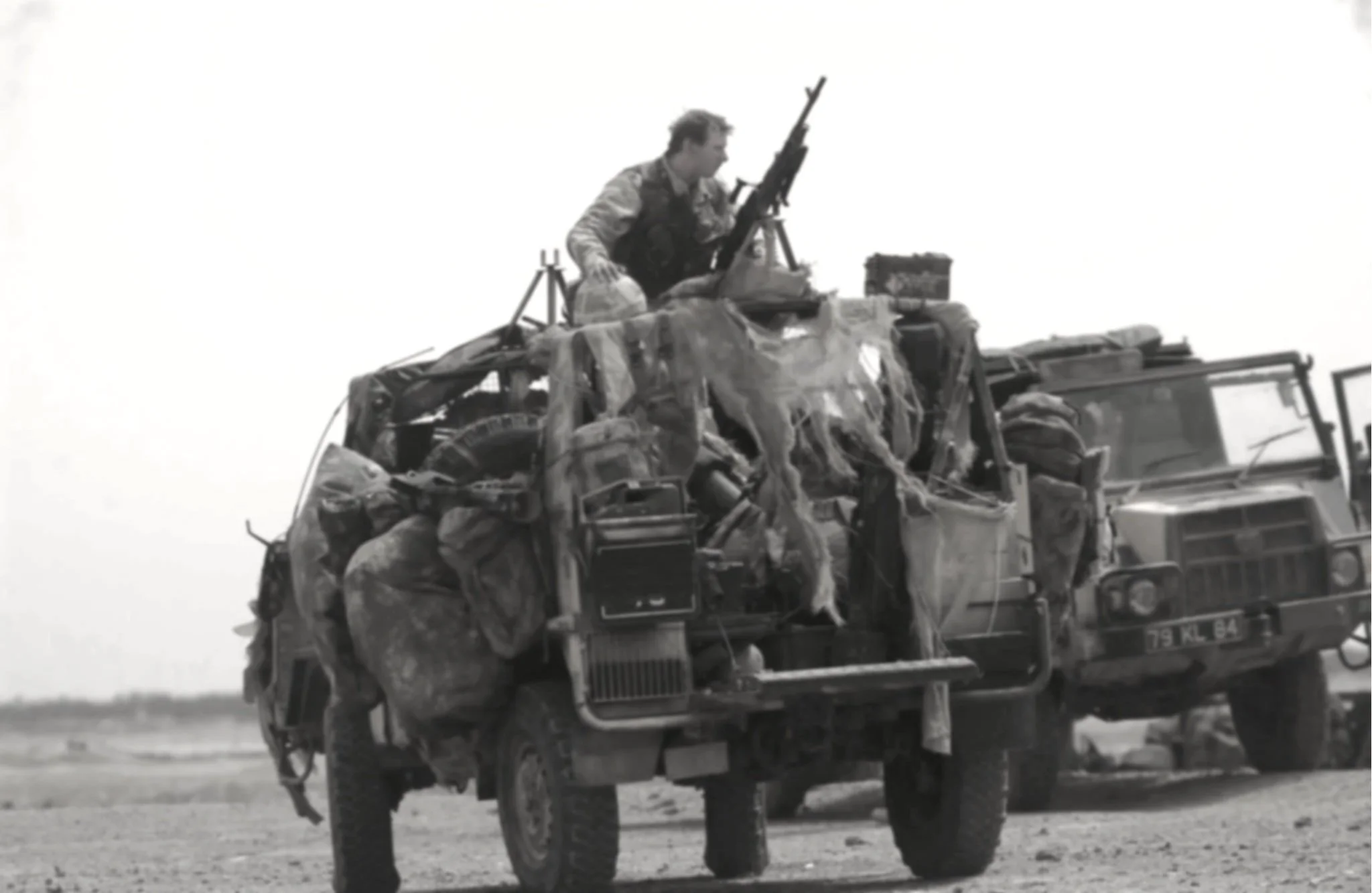A soldier looking through binoculars while sitting on top of a military vehicle, with cloudy sky in the background.