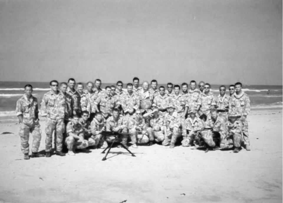 Black and white photo of a group of soldiers in camouflage uniforms on a beach, with some holding a drone and a ball.