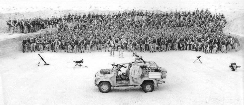 A large group of soldiers standing in formation outdoors with military equipment, including a Jeep with mounted weapons in front.