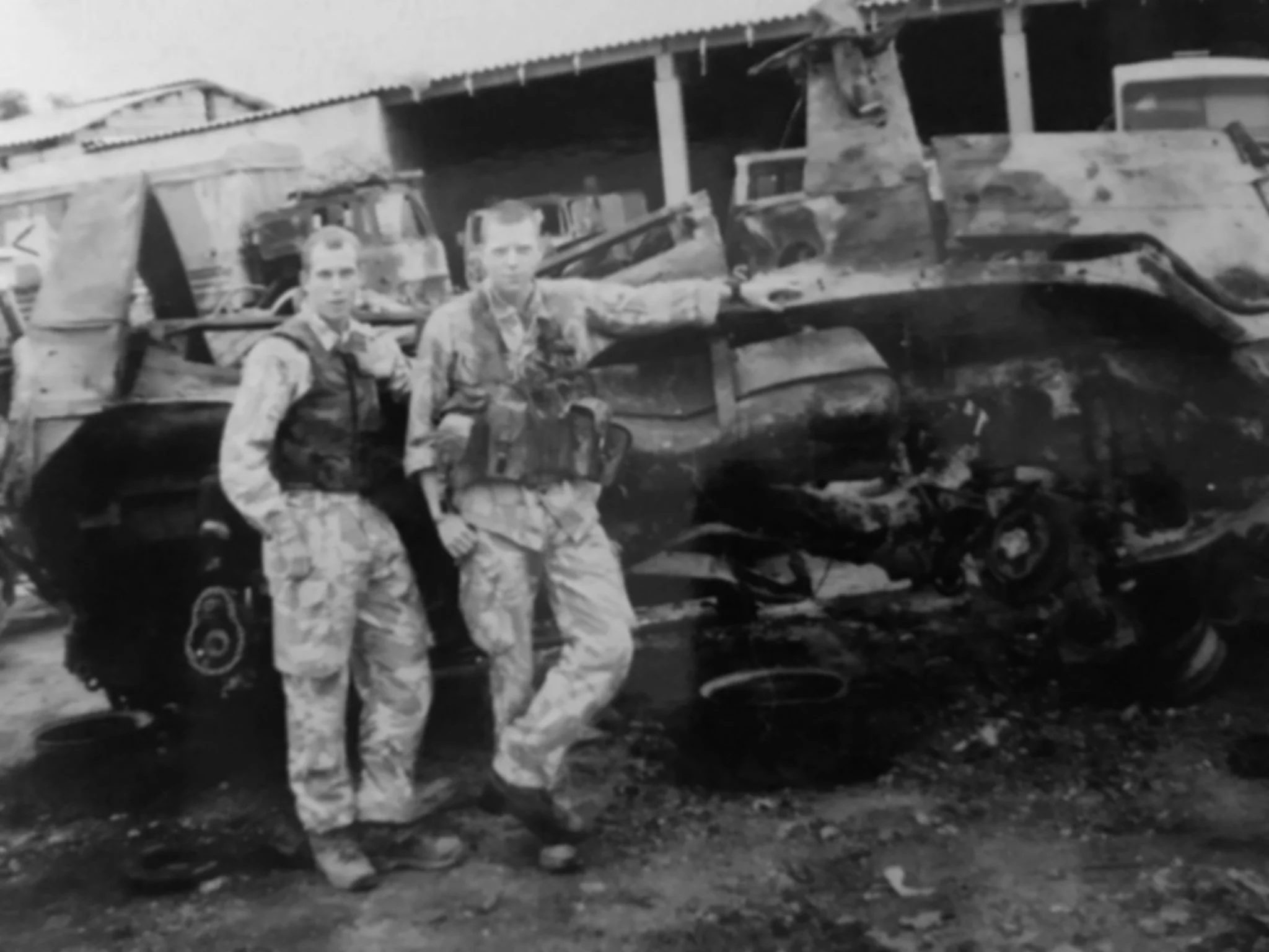 Two soldiers in camouflage uniforms and helmets standing in front of a damaged military helicopter with a burned-out exterior, parked on the ground.