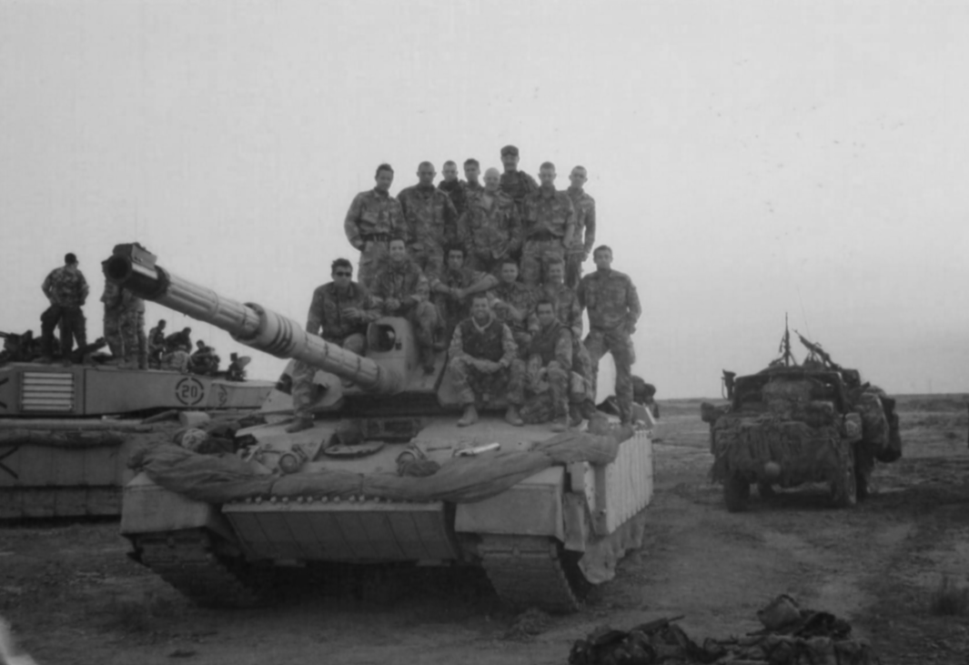 Group of soldiers sitting and standing on a tank in a barren landscape with another military vehicle nearby.