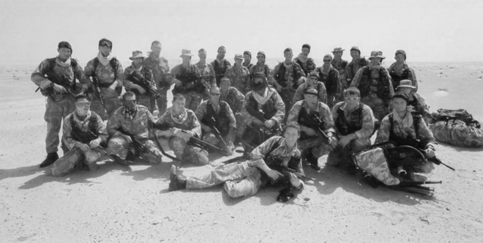 Group of soldiers in desert camouflage uniform posing with rifles on sandy terrain, some kneeling and others standing, in a desert environment.