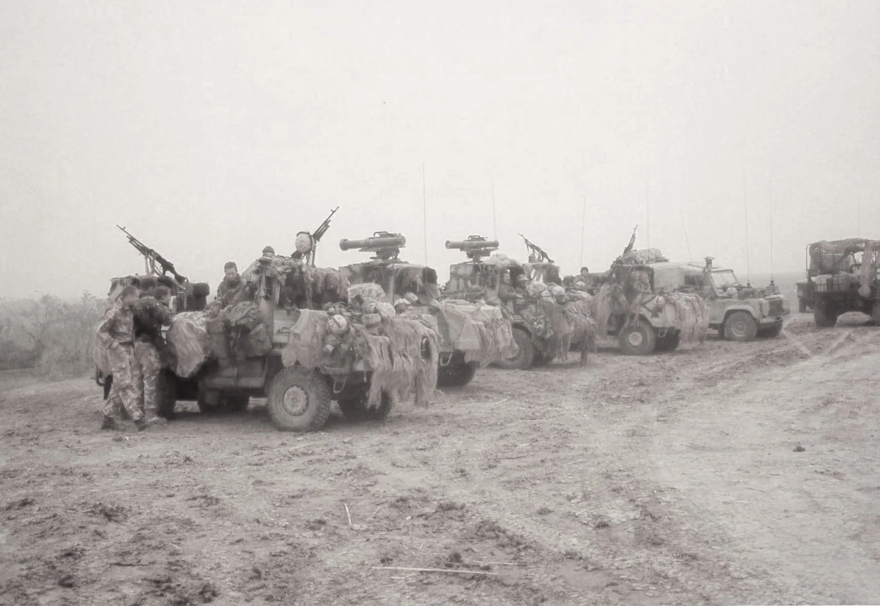 A line of military vehicles with soldiers standing around and on them in a dusty open field.