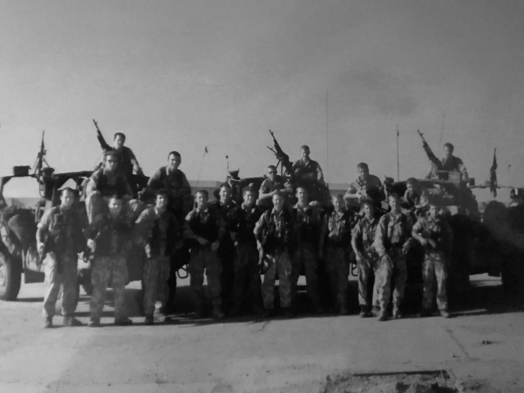 Black and white photo of a group of soldiers standing and sitting in front of military vehicles, with some soldiers standing on top of the vehicles, holding rifles.