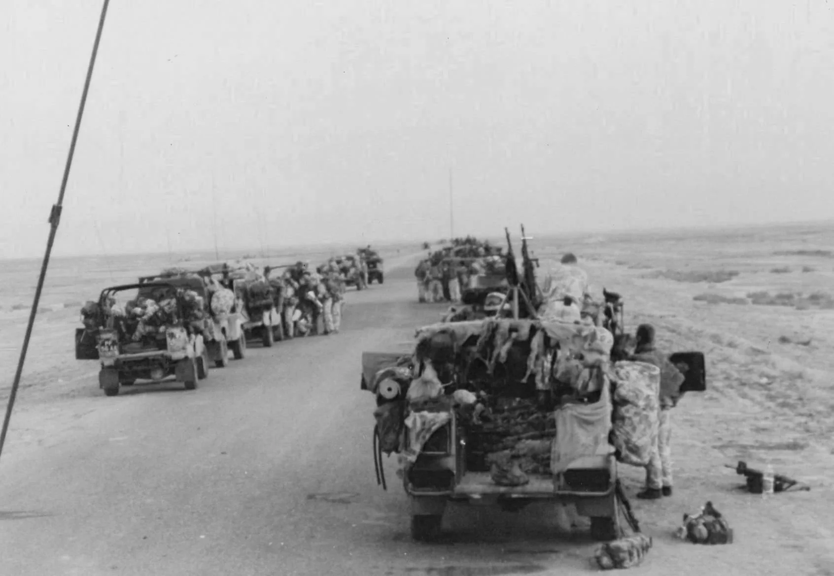 A line of military vehicles and personnel on a dirt road in an open desert landscape, with luggage and supplies on and around the vehicles.