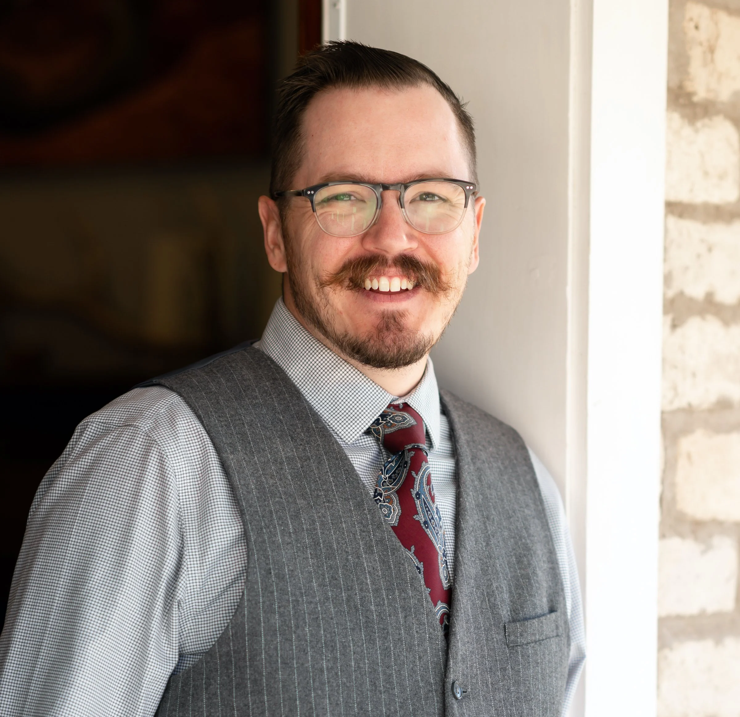 Man with glasses and mustache wearing a gray vest and tie, smiling indoors.