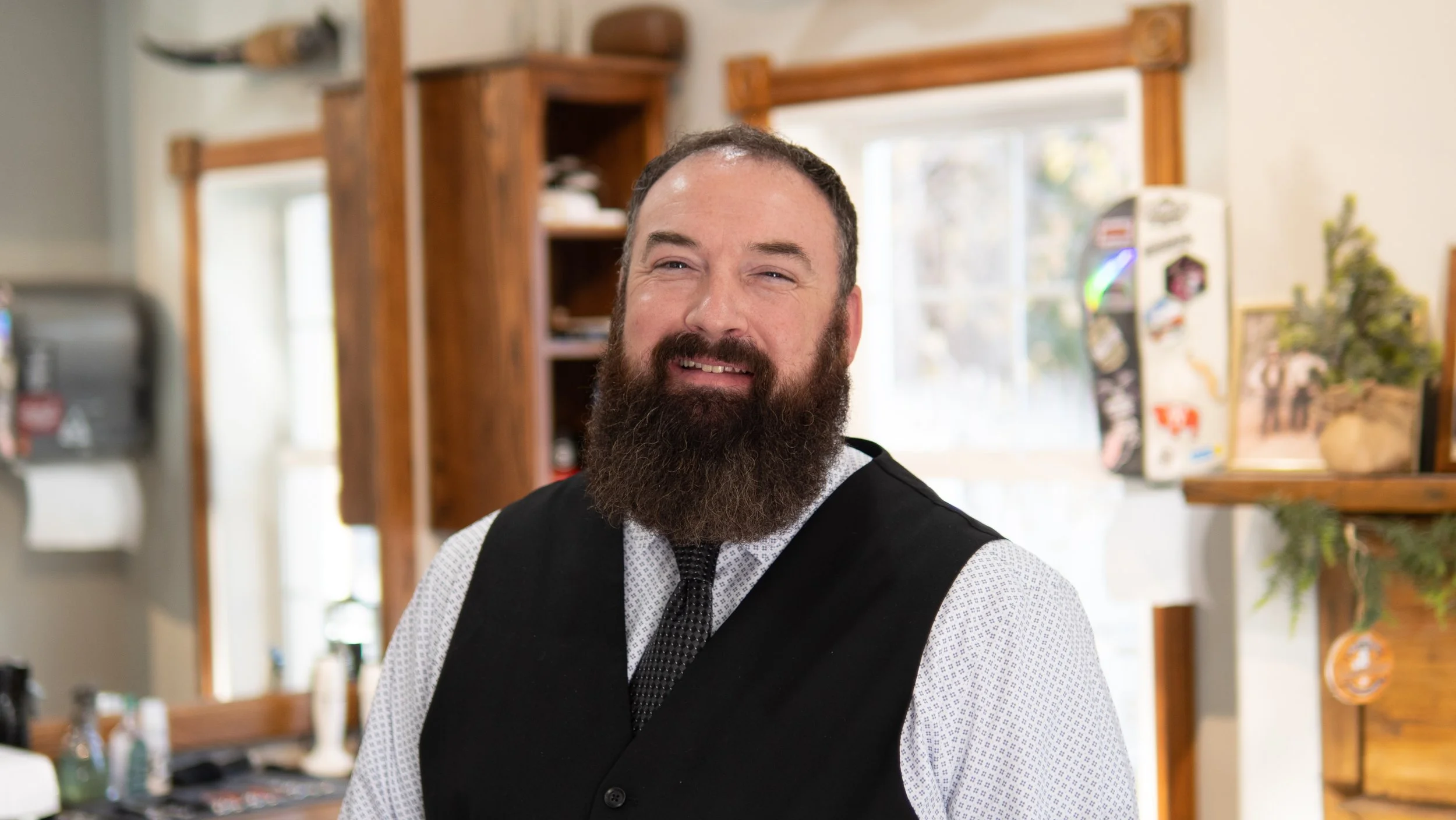 Man with a beard wearing a black vest and tie standing indoors in a rustic setting.