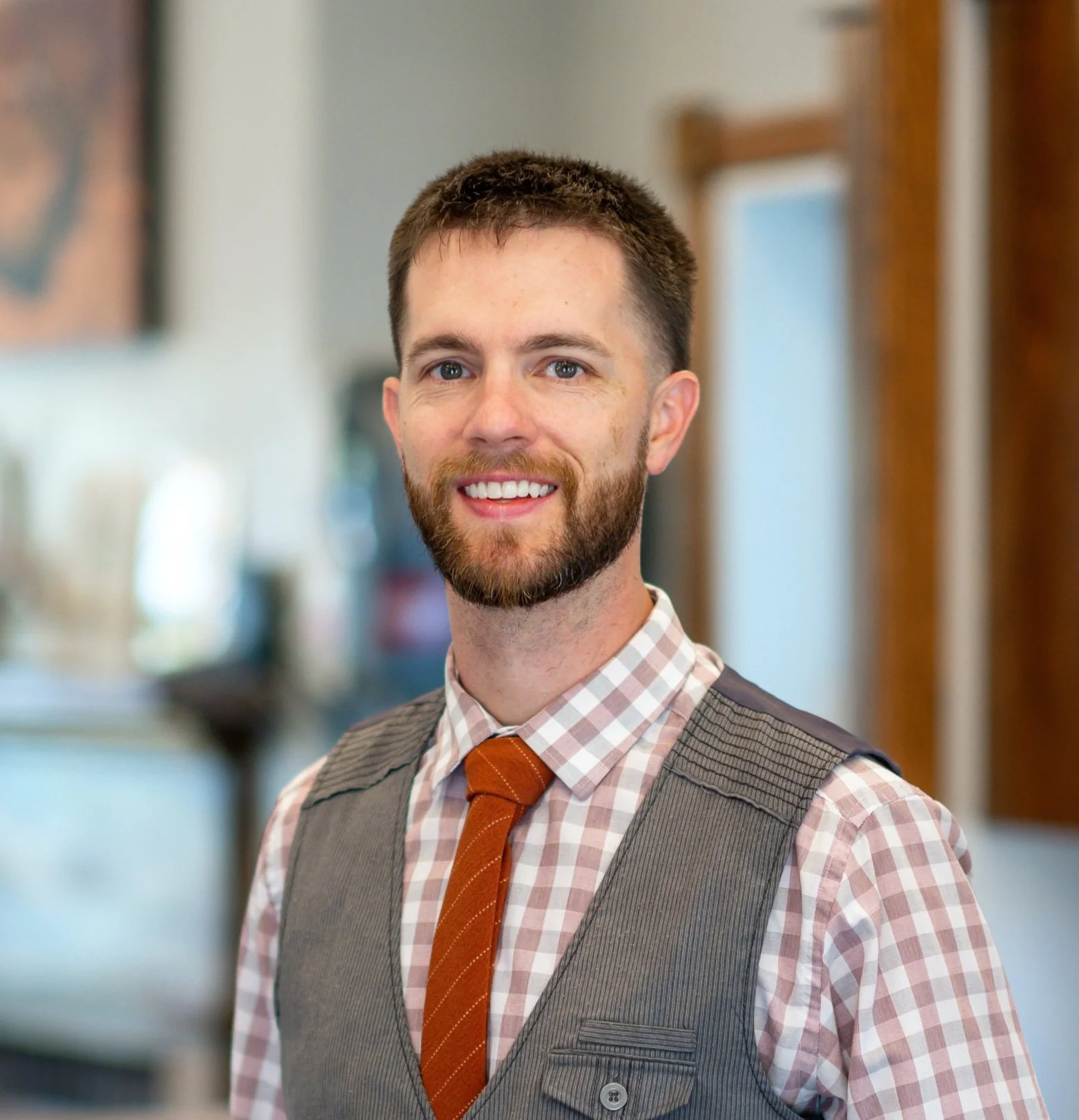 Man with a beard wearing a plaid shirt, orange tie, and gray vest smiling indoors.
