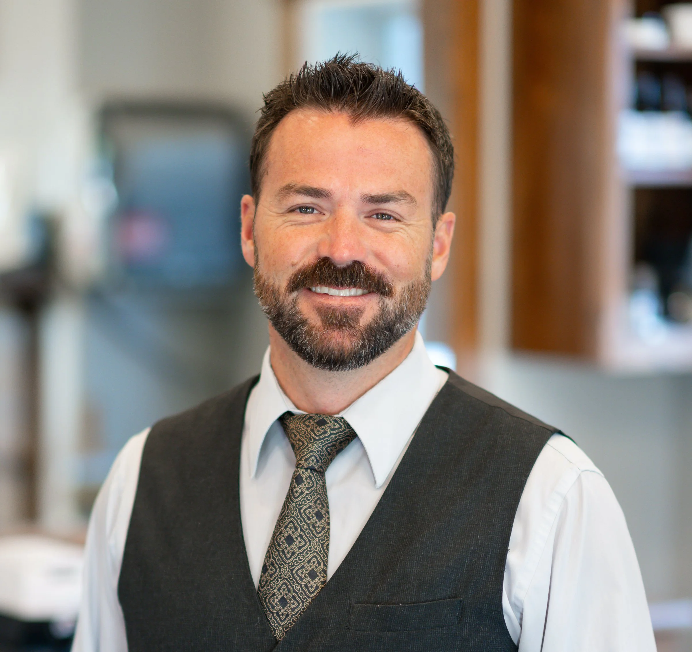Smiling man in a gray vest and patterned tie in an indoor setting.
