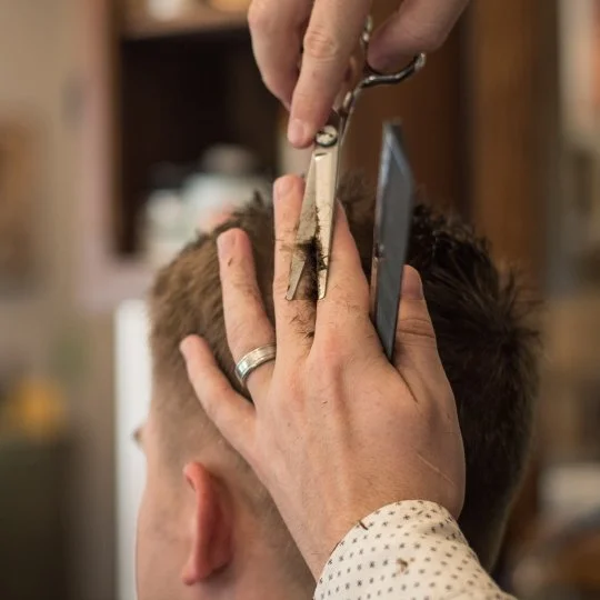 Close-up of a barbershop haircut, showing hands cutting a man's hair with scissors and a comb.