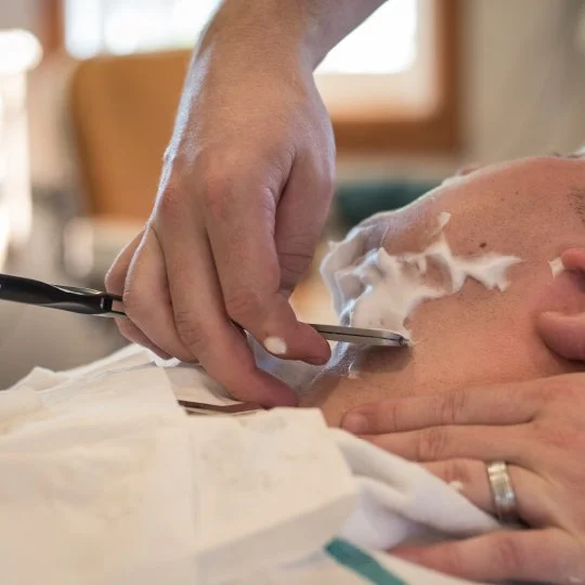 Close-up of a barber using a straight razor to shave a man's face, with shaving cream applied.