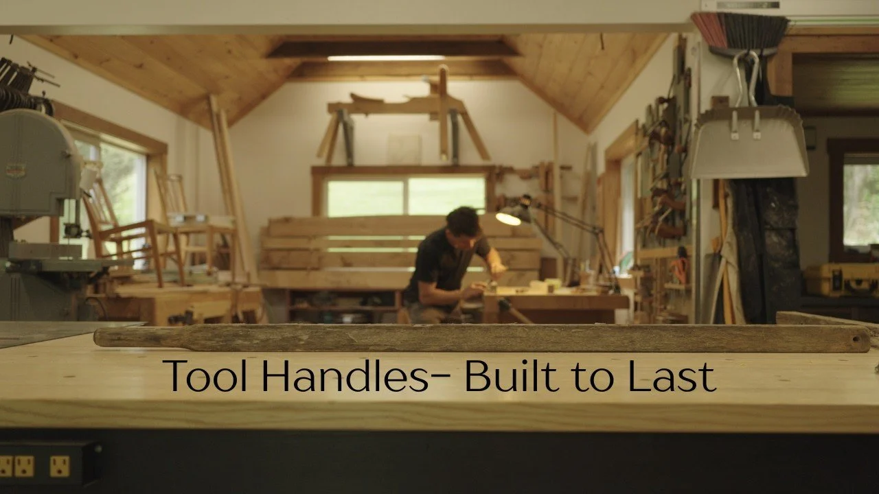 A man in a wood shop, surrounded by tools, hand crafts new handles for a wheelbarrow from native wood.
