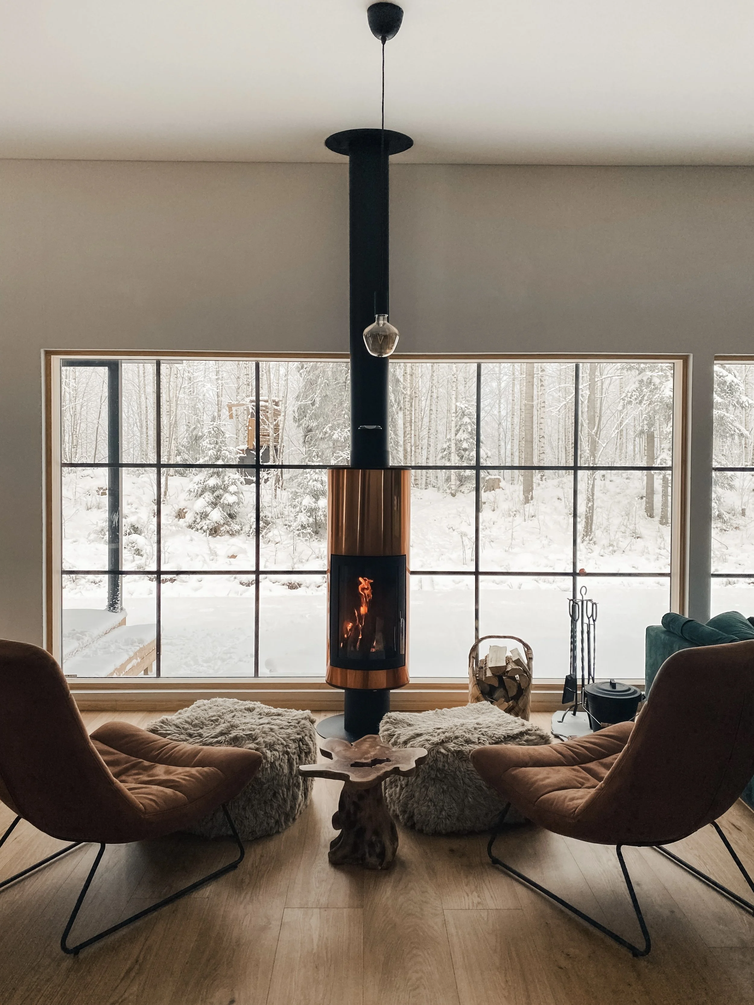 Sisäkuva olohuoneesta, jossa on takka ja kaksi nojatuolia, jotka on päällystetty ruskealla sametilla. Takan vieressä on iso ikkuna, josta näkyy luminen metsämaisema. Keskellä huonetta on uniikki puinen pöytä ja kaksi harvinaisen pehmeää talja-tyynyä. Olohuoneessa on myös muita huonekaluja ja koriste-esineitä, ja huoneessa on lämpöä ja kodikkuutta.