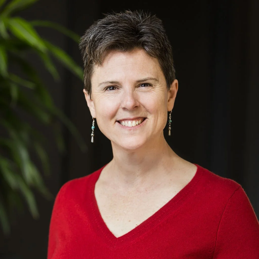 Portrait of a smiling woman with short dark hair, wearing a red V-neck top and dangling earrings, standing in front of a dark background with a green plant visible on the side.