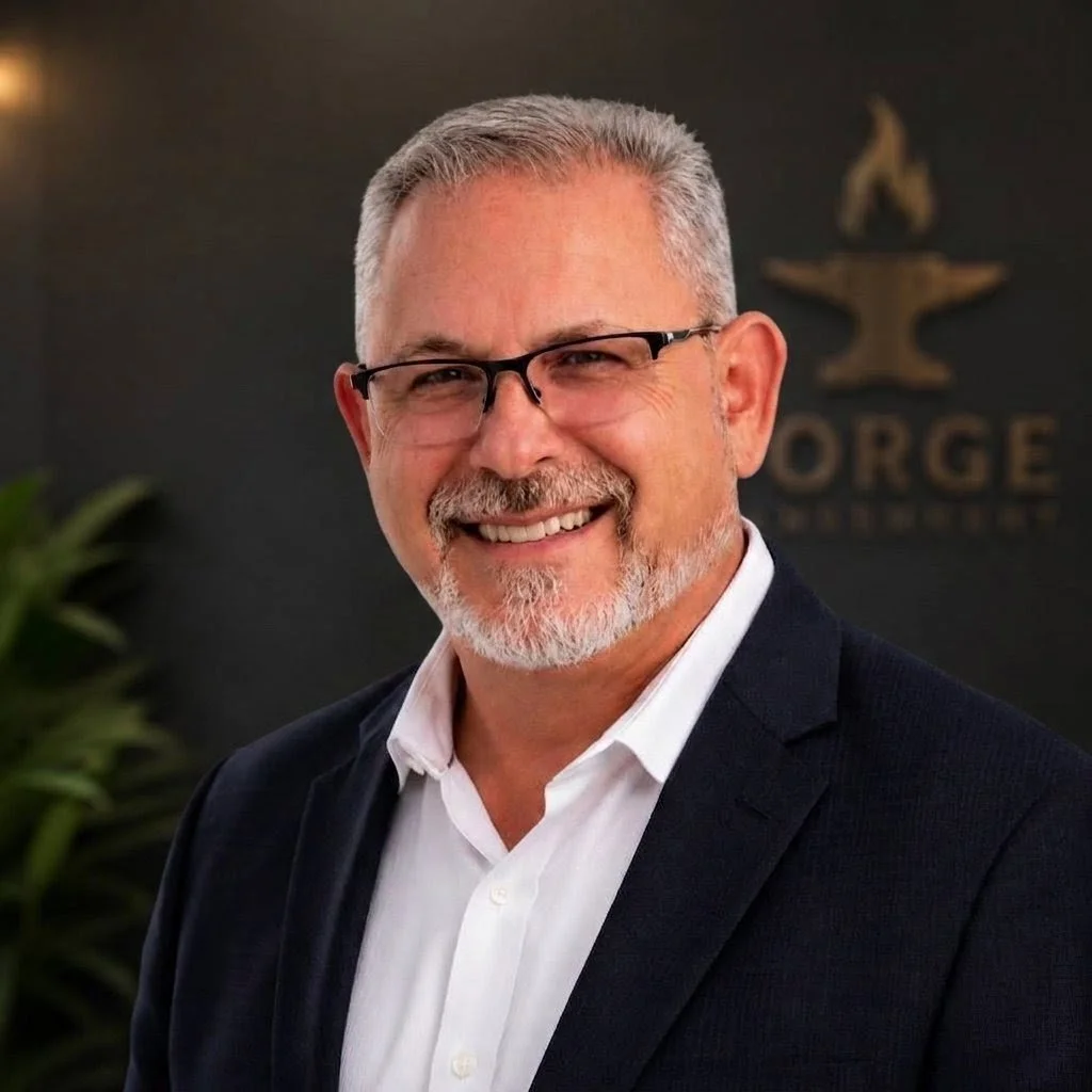 A smiling middle-aged man with short gray hair and glasses, wearing a dark suit jacket and white shirt, standing indoors in front of a blurred sign with the words 'Office' visible.