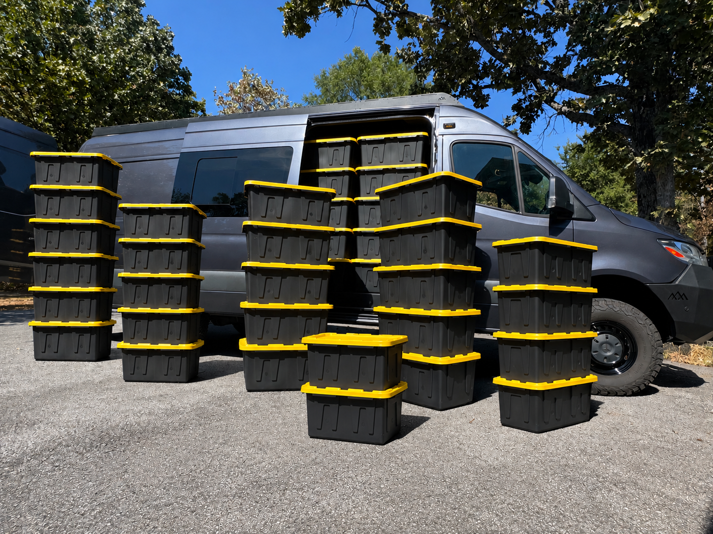 A gray delivery van parked outdoors with numerous black storage bins with yellow lids arranged in front of it, on a paved surface with trees and blue sky in the background.