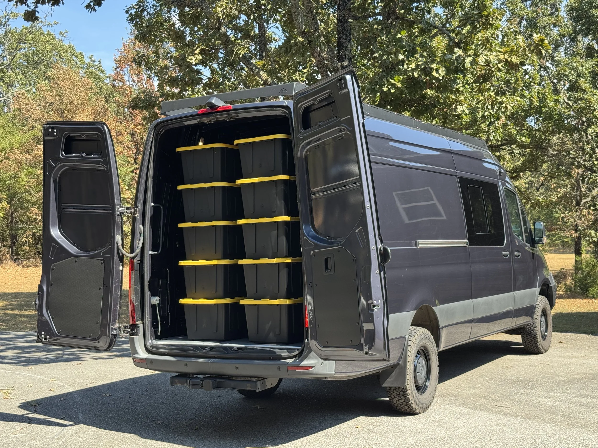 Open rear doors of a black cargo van with shelves containing black storage bins outside in a parking lot surrounded by trees