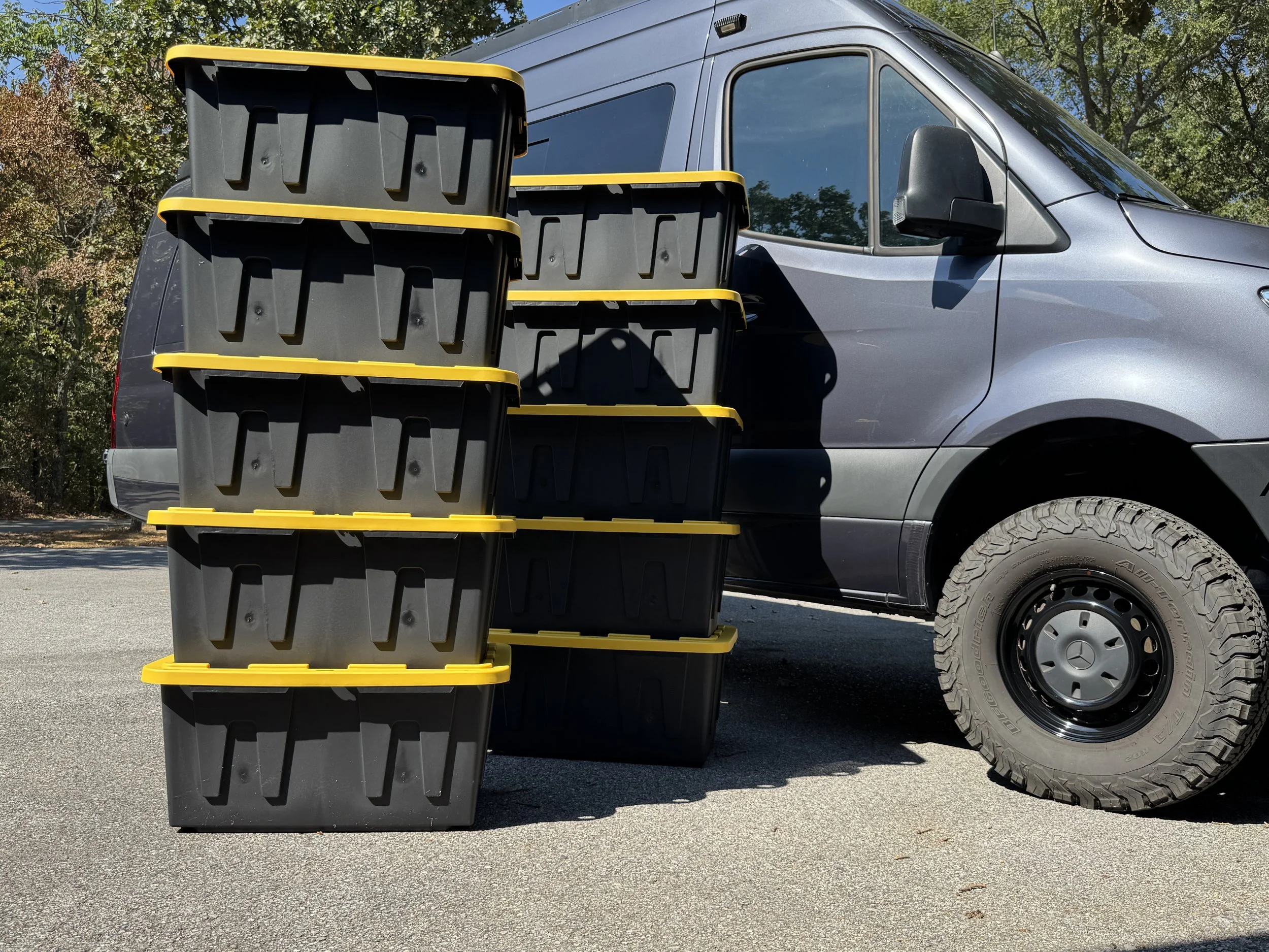 Stack of black plastic storage bins with yellow lids in front of a gray van with large tires, outdoors on a sunny day, with trees in the background.
