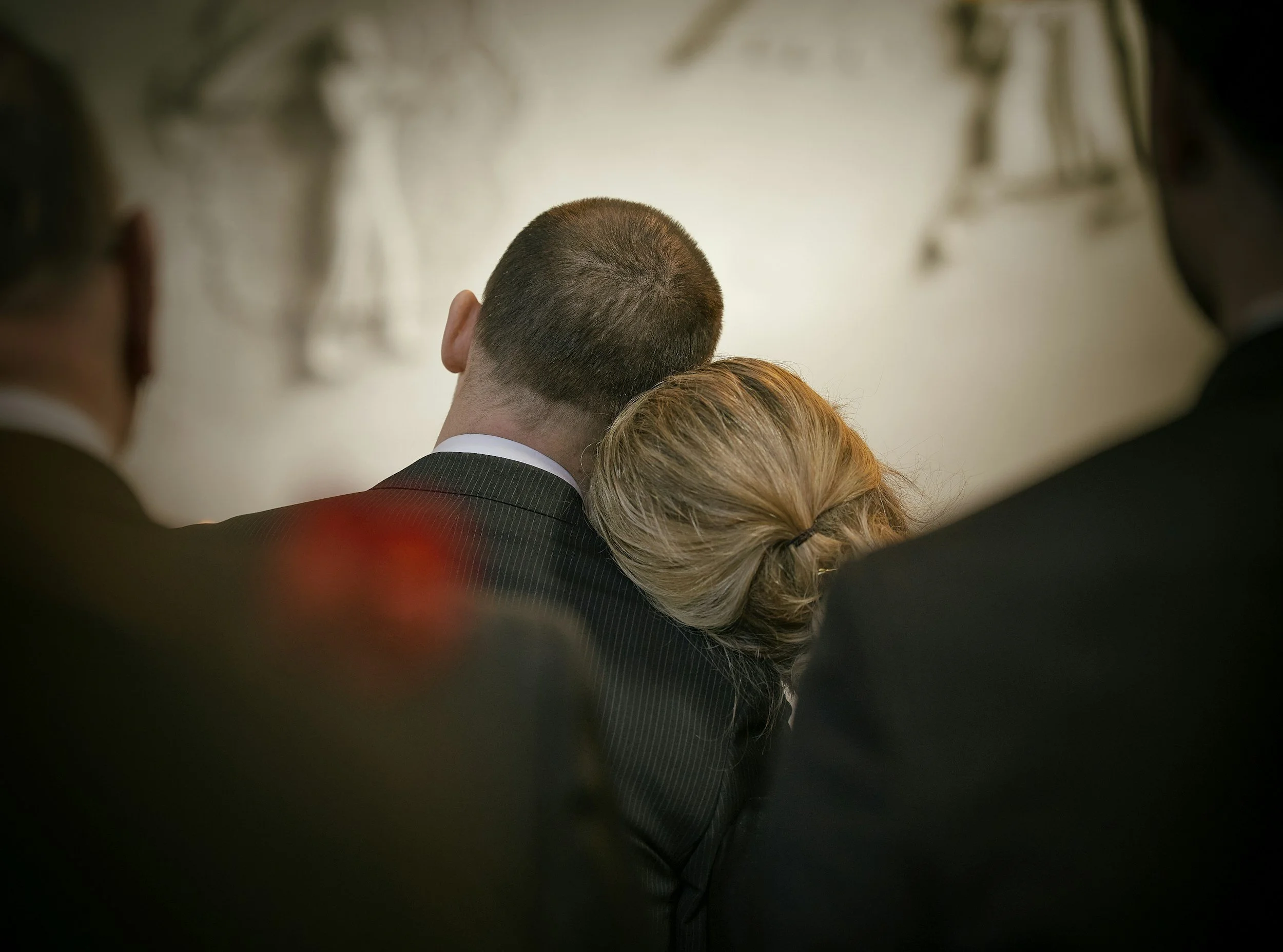 Rear view of a man with short brown hair wearing a pinstripe suit, surrounded by other people, at an indoor event.