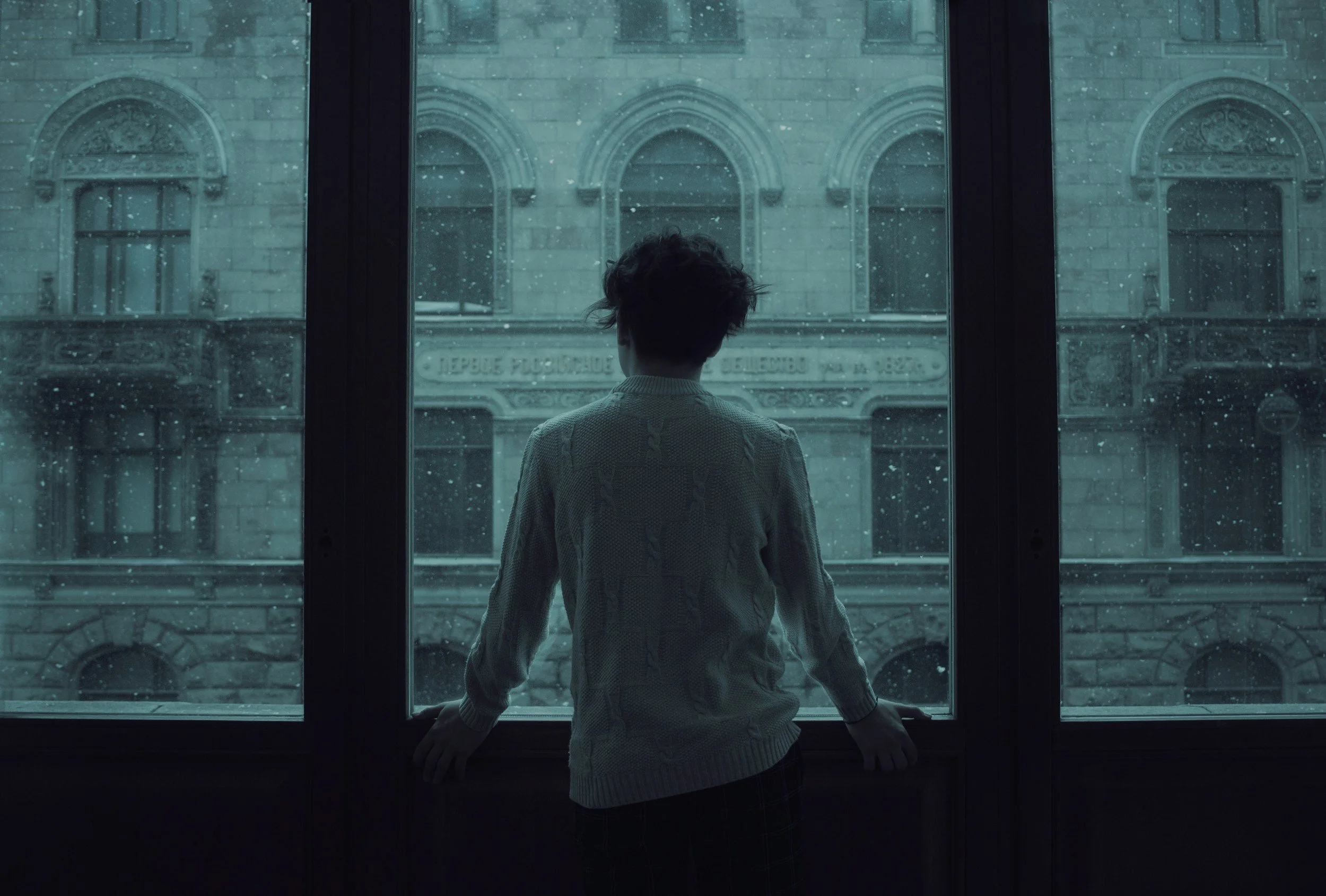 Person with curly hair wearing a sweater, standing by a window and looking outside on a rainy day with an old building visible across the street.