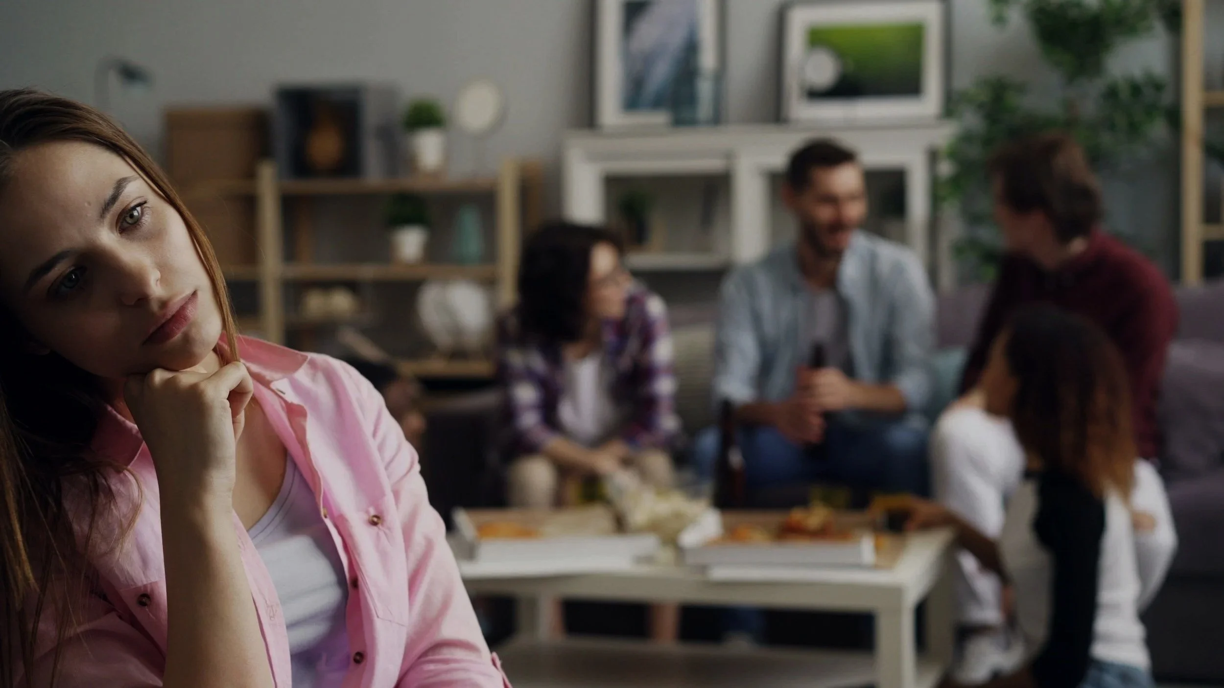 A young woman with red hair, wearing a pink jacket, thinking or contemplating in a room with a blurred group of people in the background.