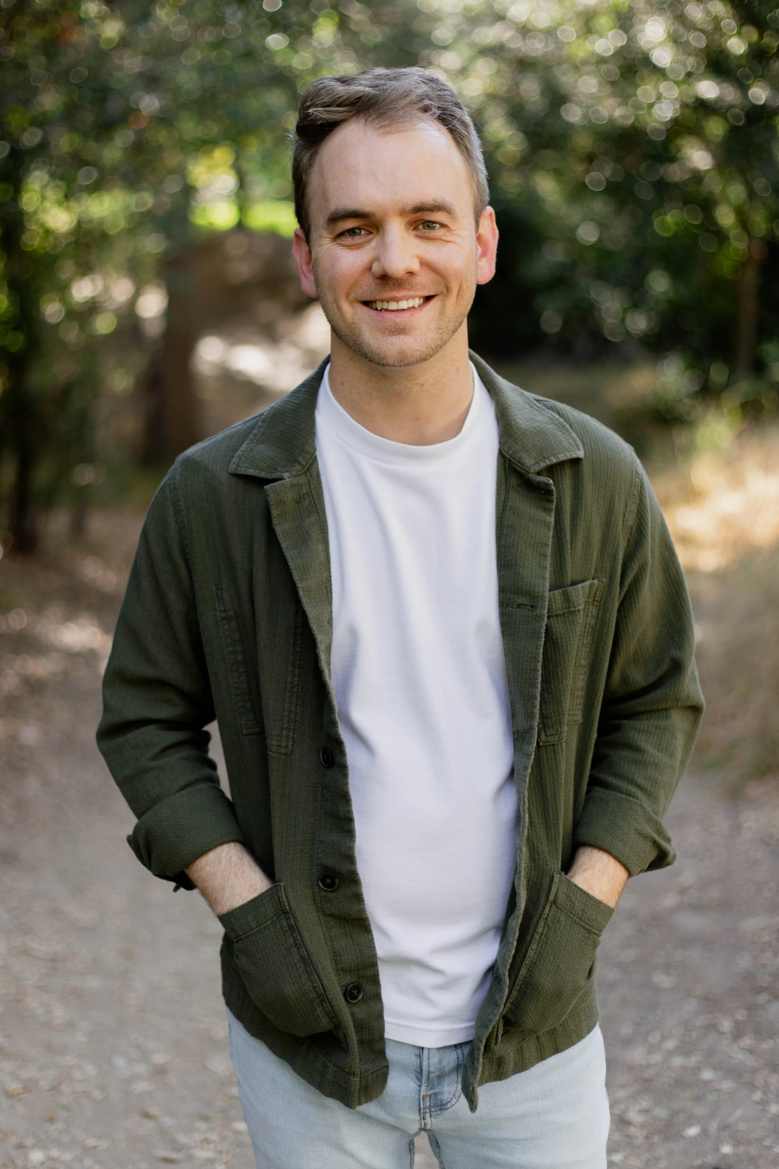 A smiling young man with short brown hair, wearing a white t-shirt and an unbuttoned green jacket with rolled-up sleeves, standing outdoors on a dirt path surrounded by blurred green foliage.