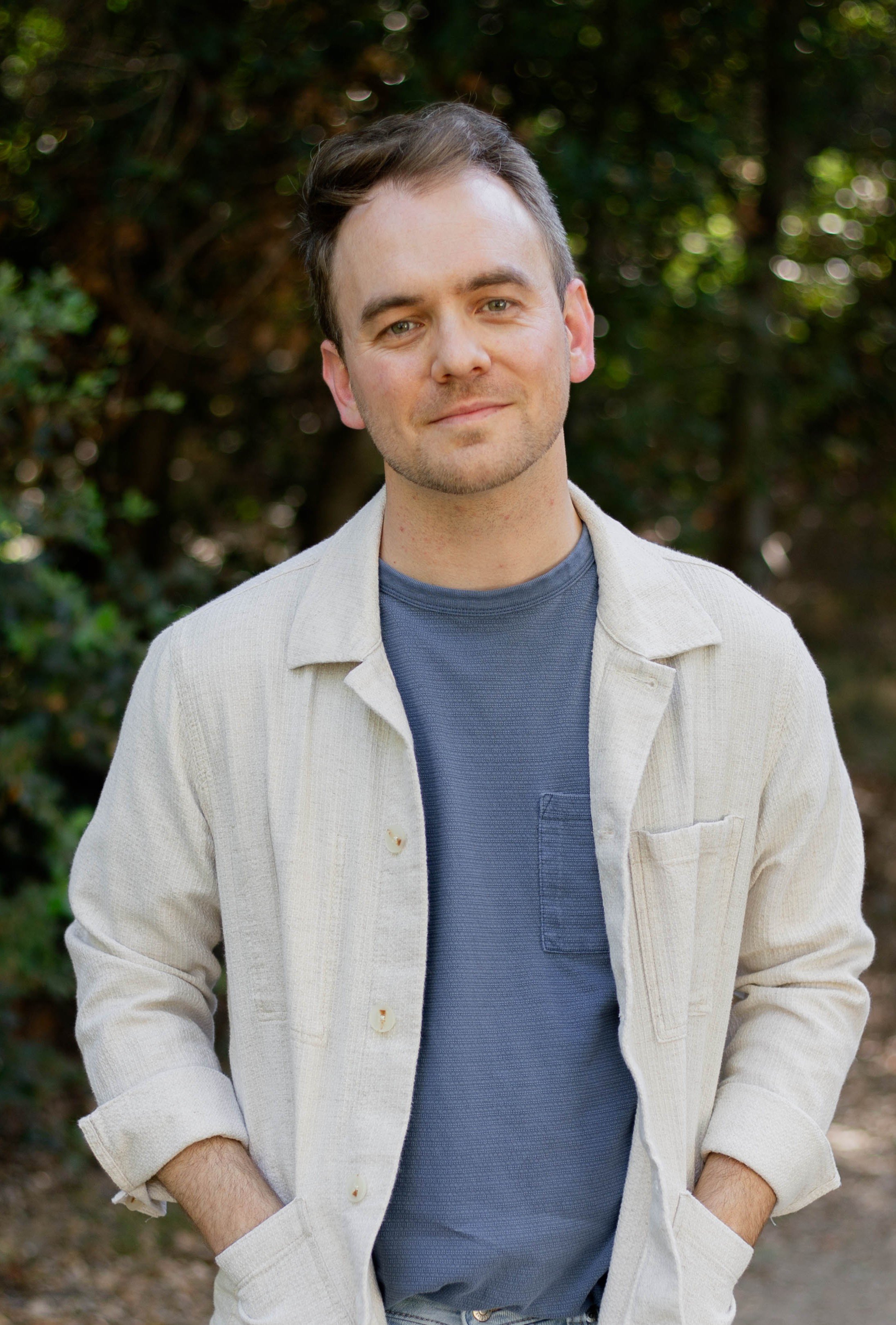 A young man with light brown hair and a light beard, wearing a beige jacket over a blue shirt, standing outdoors with a blurred green forest background.